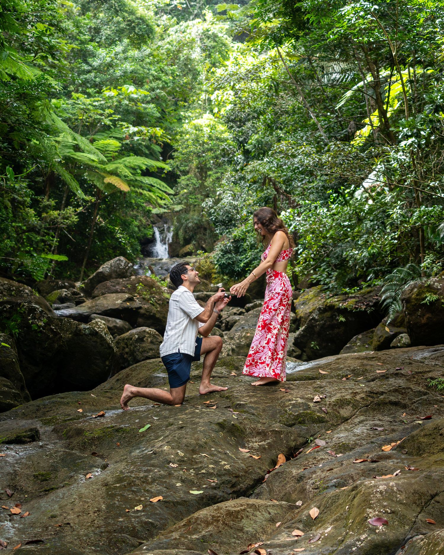 Two people climb on large rocks near a waterfall in a lush forest setting. Surprise proposal Engagement photography outside El Yunque