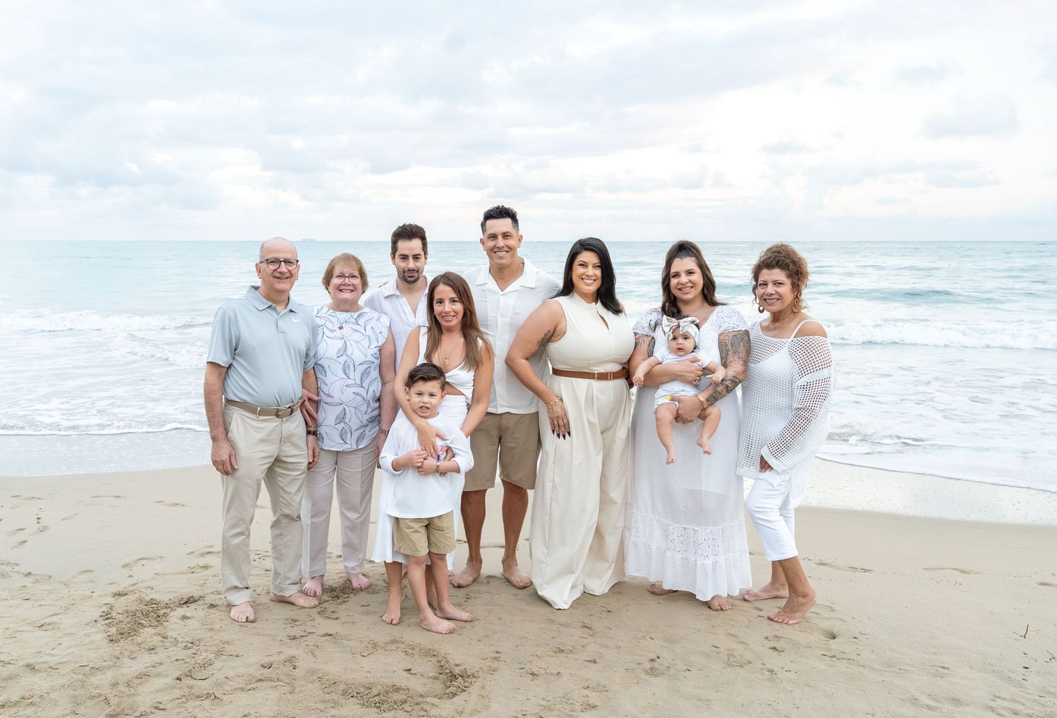 Multi-generational family in white outfits posing together on a sandy beach with ocean waves in the background.