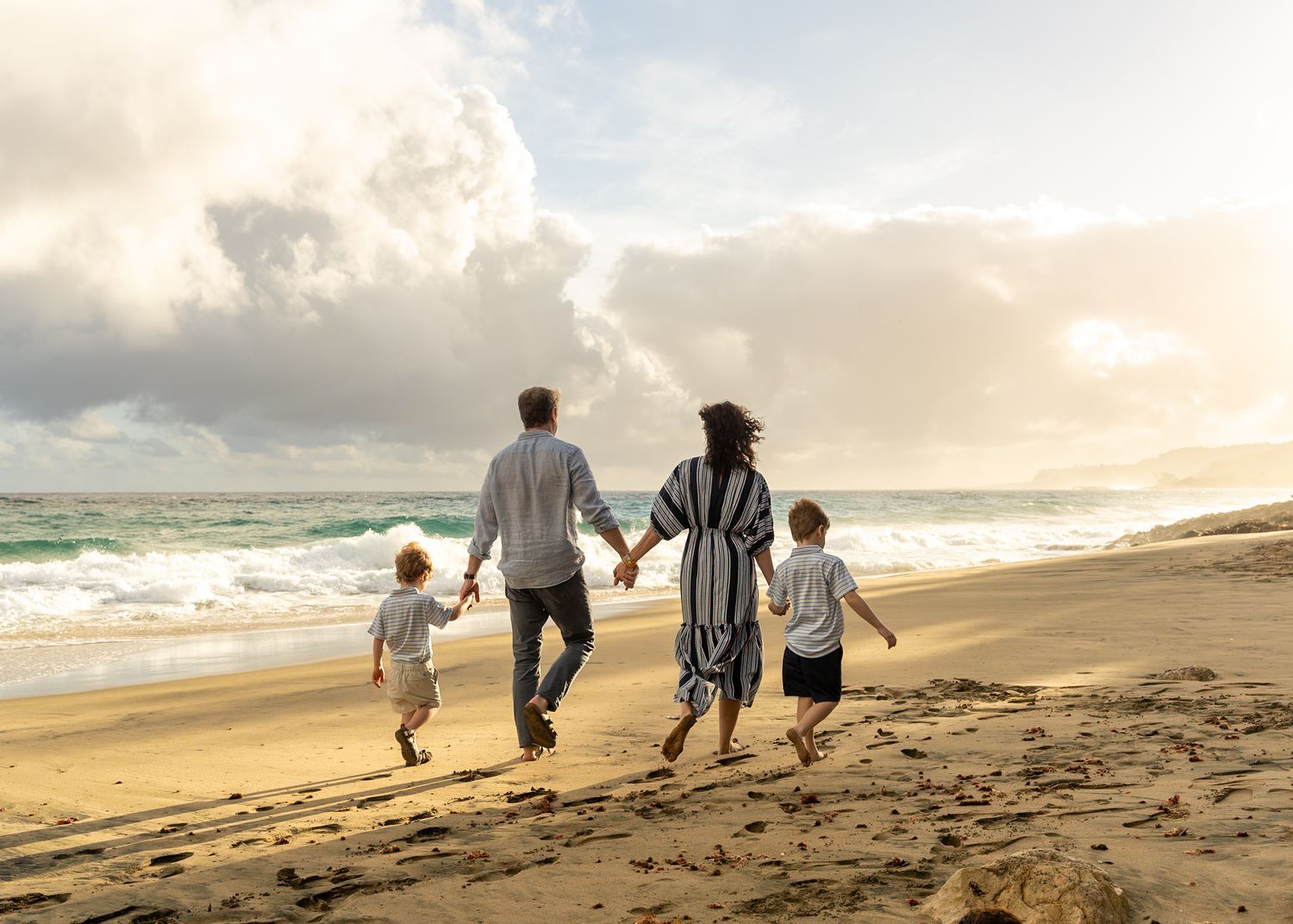 Family of four walking hand-in-hand along a sandy beach with ocean waves and dramatic cloudy sky.