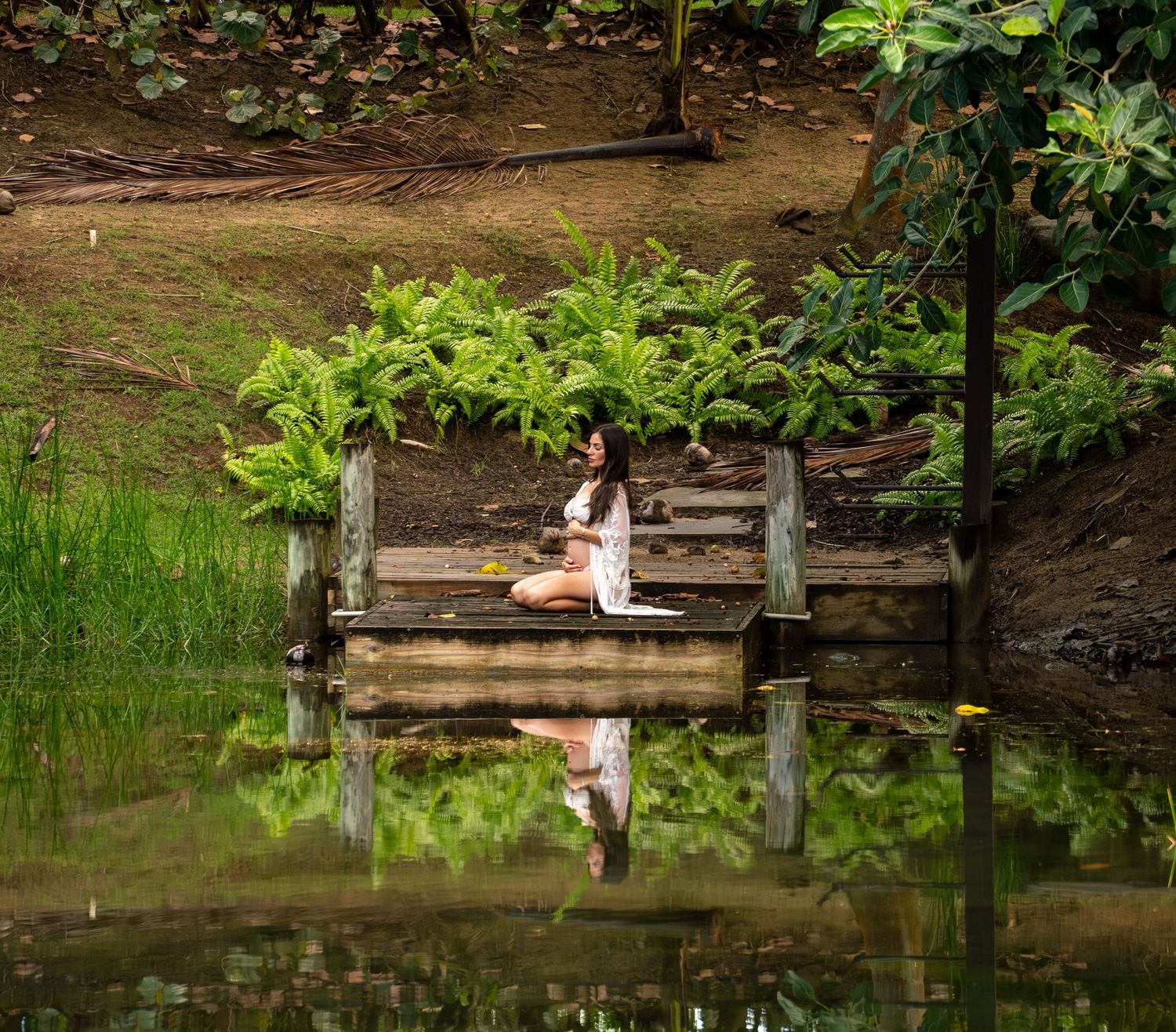 Woman meditating on a wooden dock surrounded by lush green ferns and calm reflective water in a serene garden.