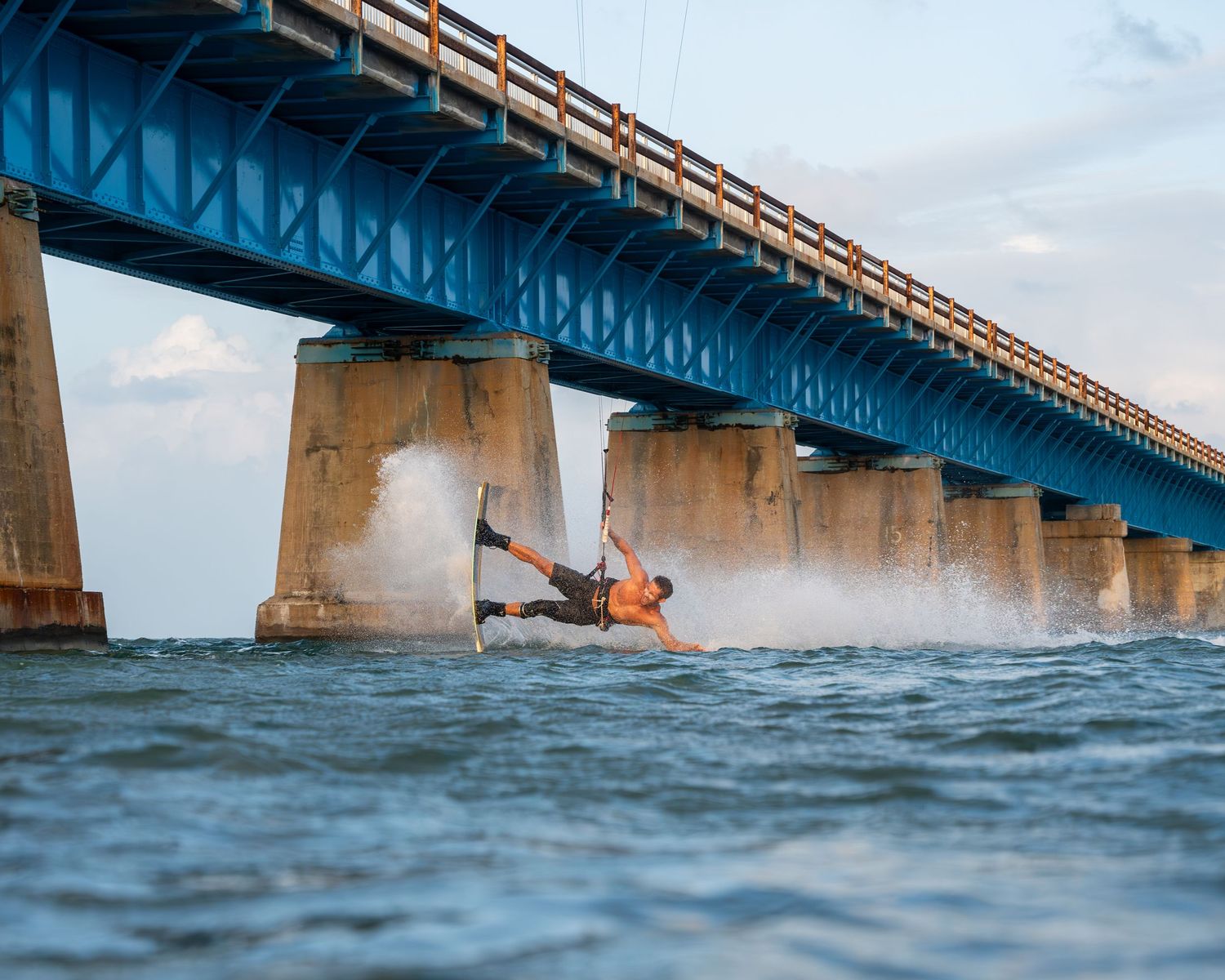 Kitesurfer riding waves near a large blue bridge structure over ocean waters.