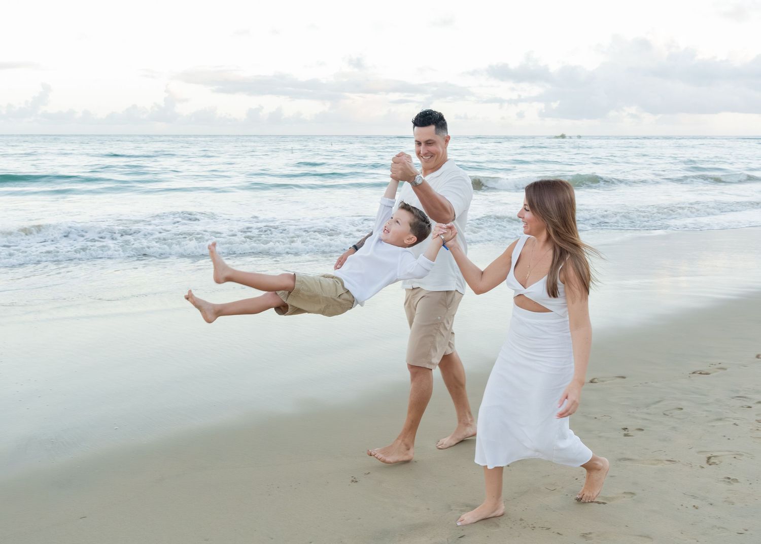 Family in white outfits playfully swinging child on a tropical beach with turquoise waves in the background.