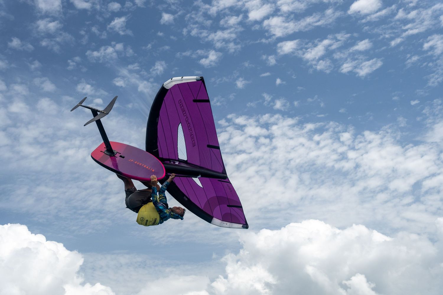 Purple kiteboarding wing silhouetted against a cloudy blue sky during an aerial maneuver.