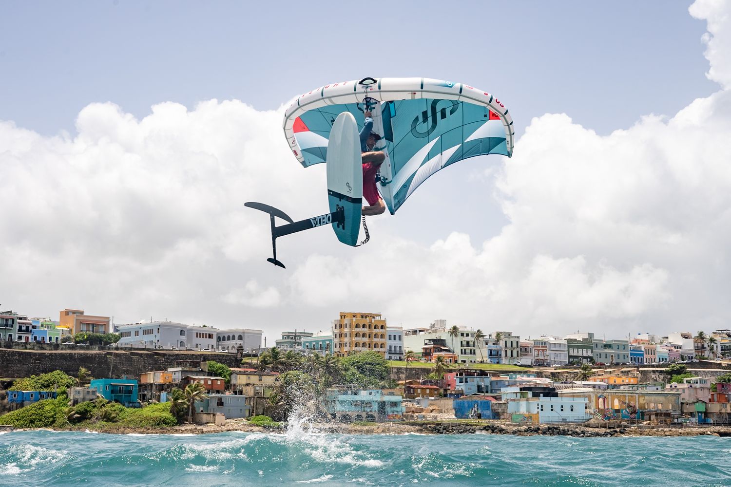 Aerial view of colorful coastal town with wing foiler riding waves in tropical waters.