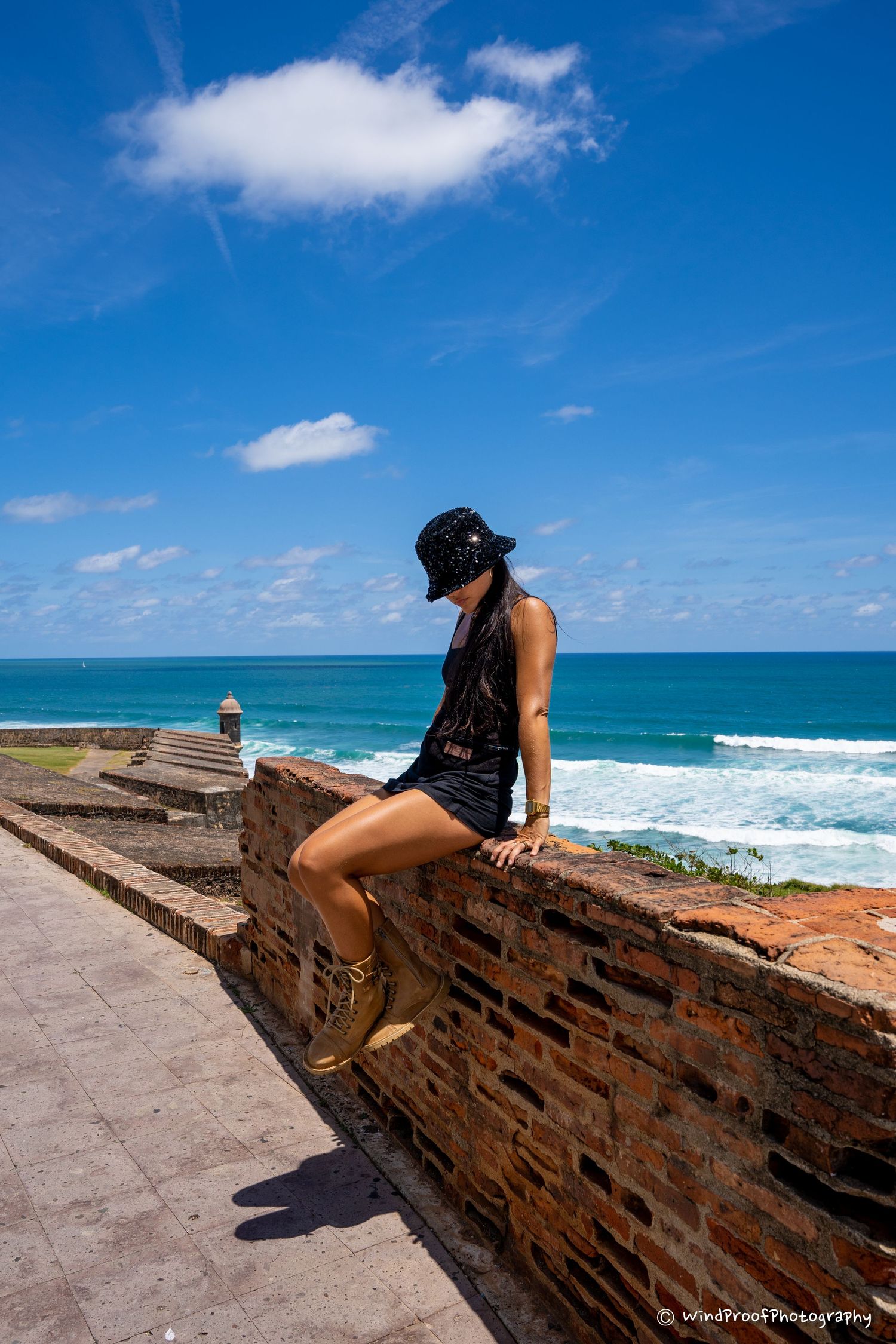 A person in casual attire sits on an oceanside stone wall overlooking turquoise waters on a sunny day.