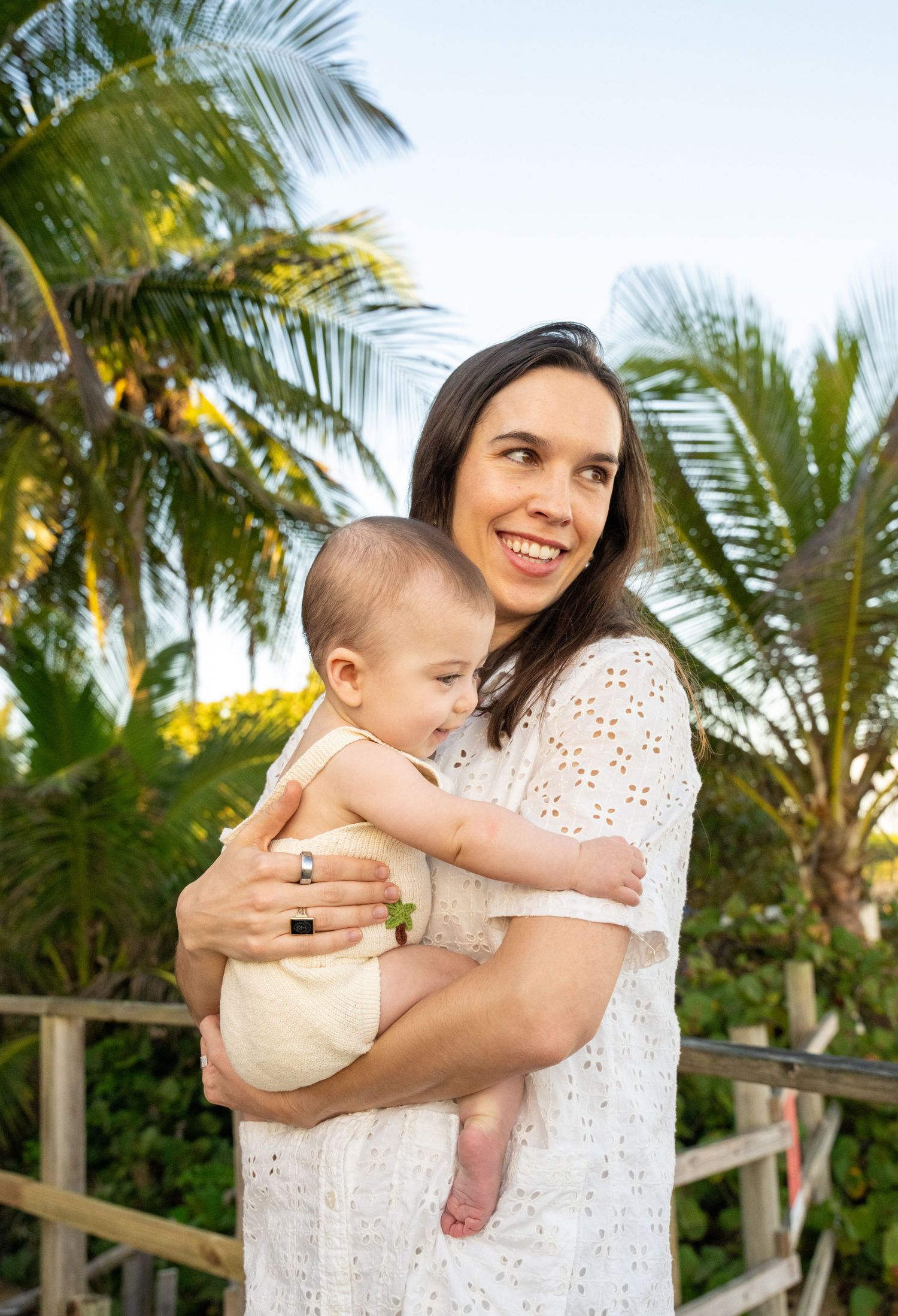 Smiling woman in white lace dress holding a baby outdoors with tropical palm trees in the background.