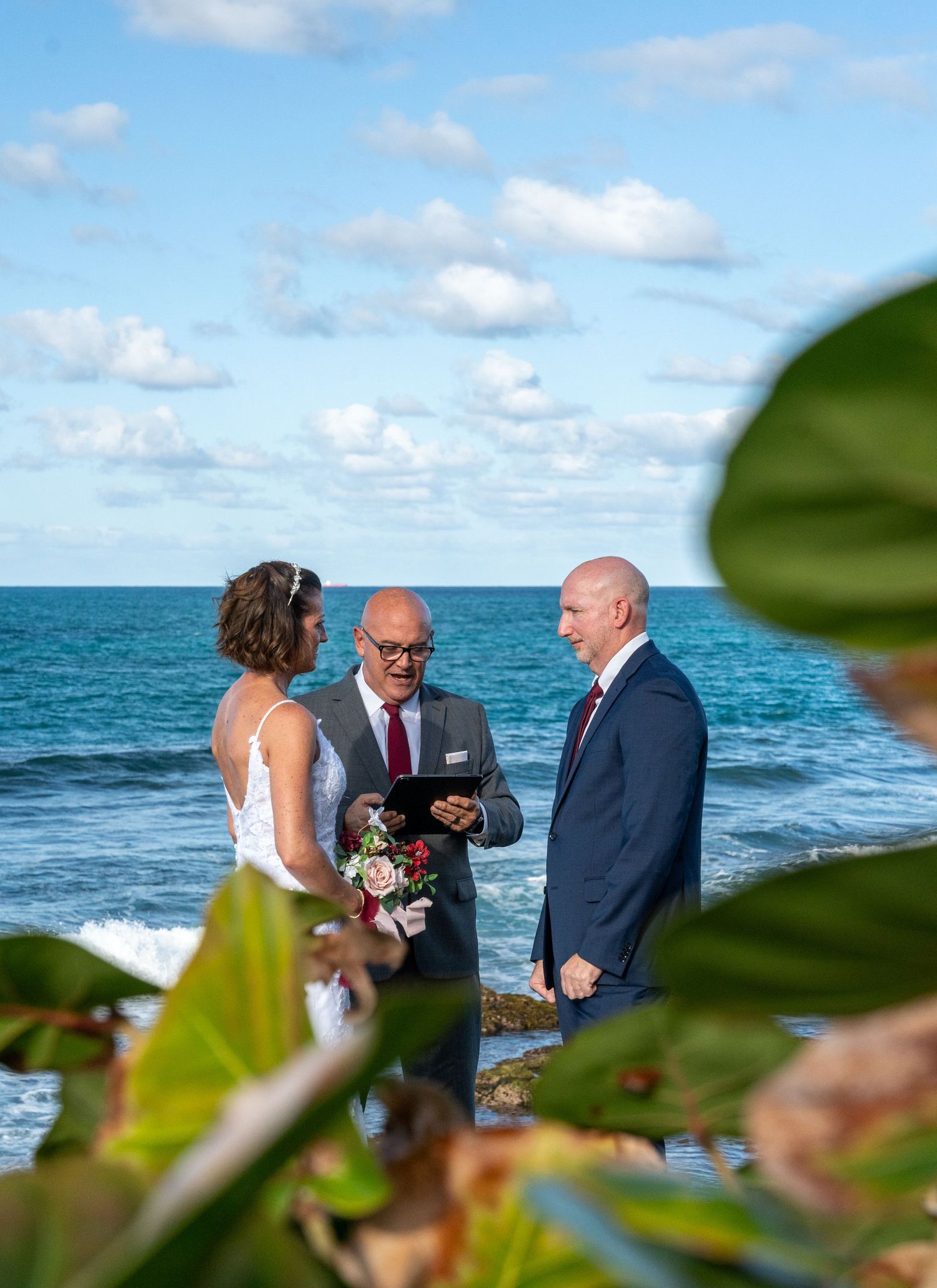 A wedding ceremony takes place by the ocean with tropical foliage in the foreground.