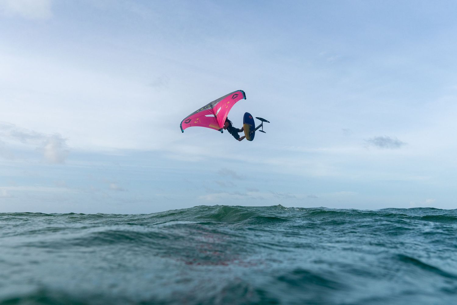 A windsurfer with a pink sail catches air above ocean waves.