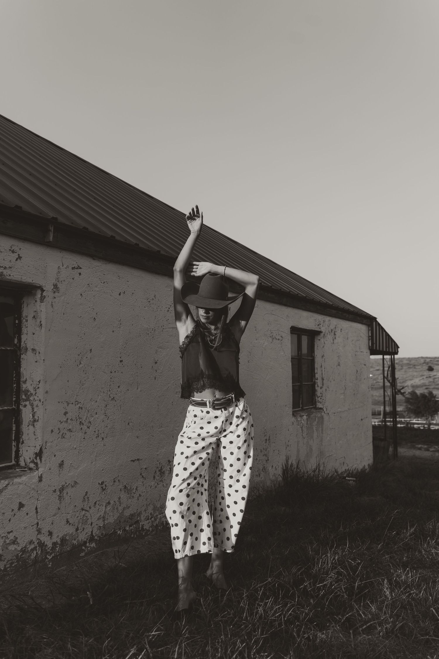 Black and white photo of a woman in polka dot pants and hat posing outdoors near a rustic building.
