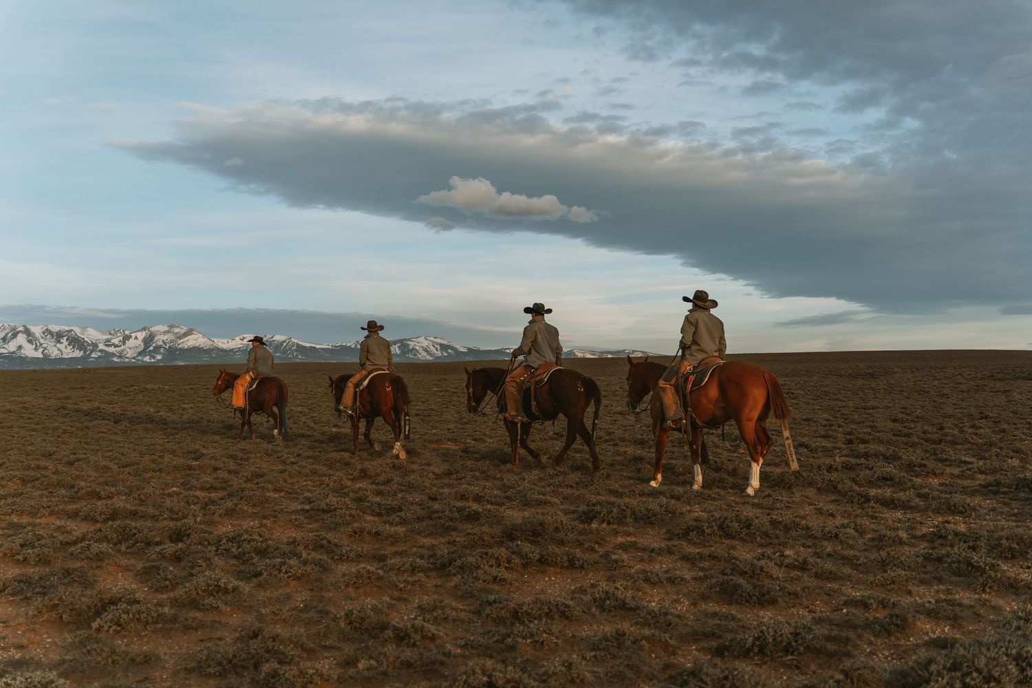 Three cowboys on horseback riding across open plains at sunset with mountain range in background.