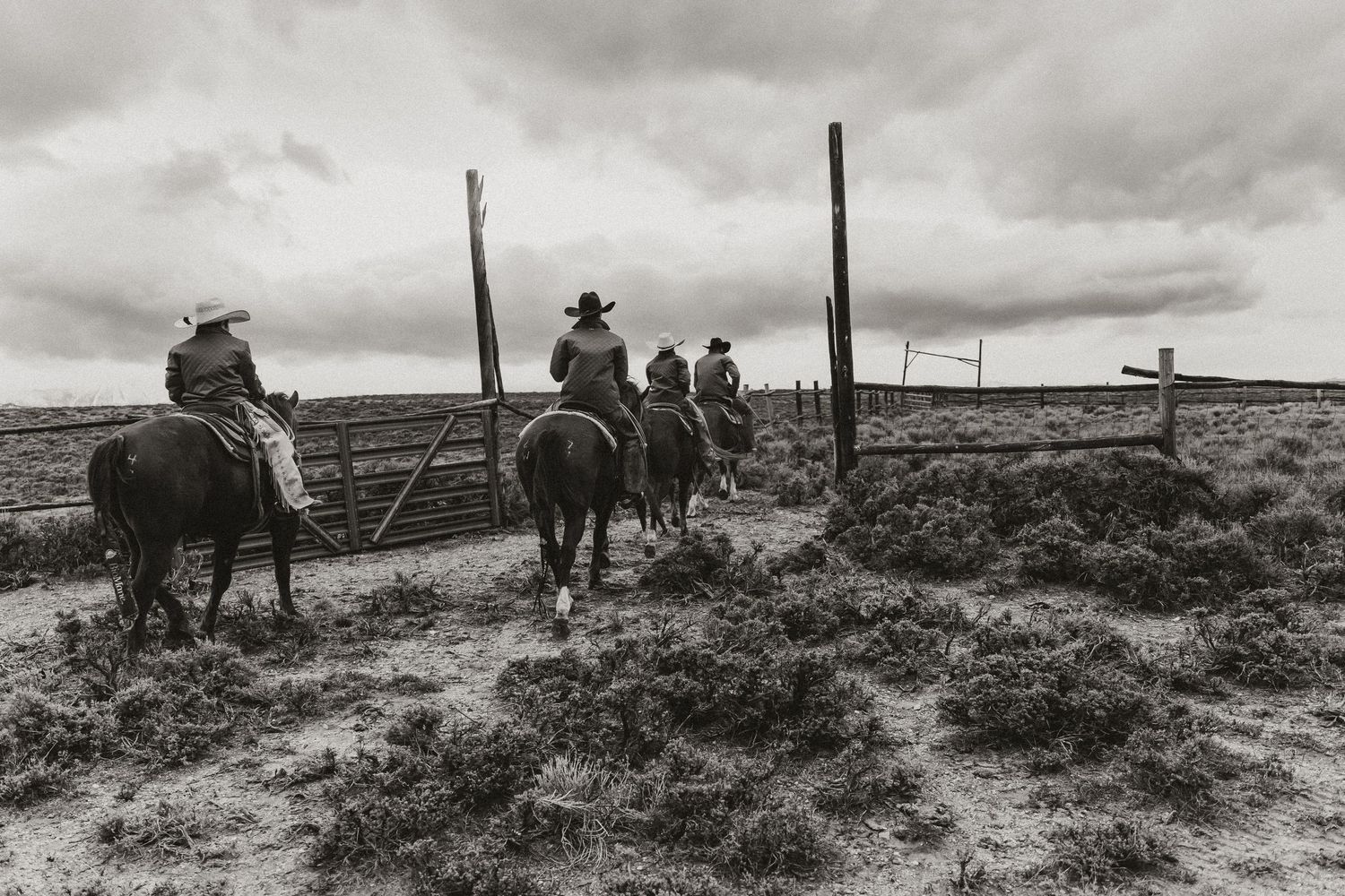 Cowboys on horseback ride through a rustic ranch gate on an open range under dramatic cloudy skies in black and white.