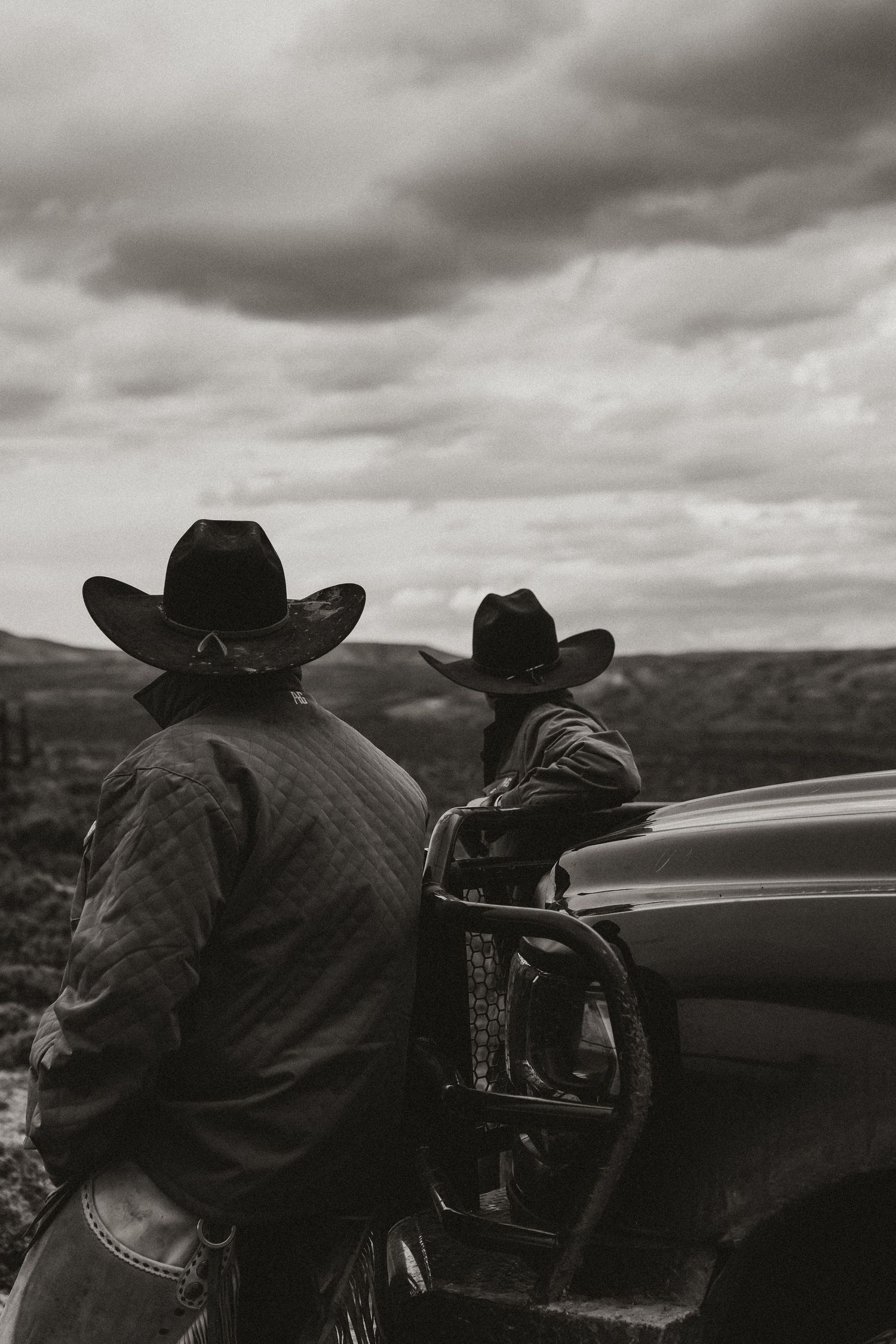 Two cowboys in black hats stand by a truck overlooking a vast landscape under cloudy skies in black and white.