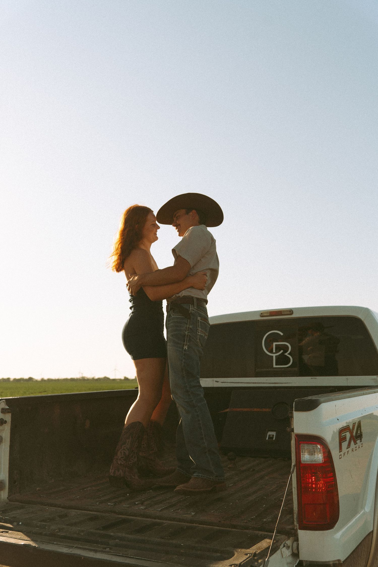 Couple embracing on pickup truck bed at sunset, woman in black dress and boots, man in cowboy hat and jeans.