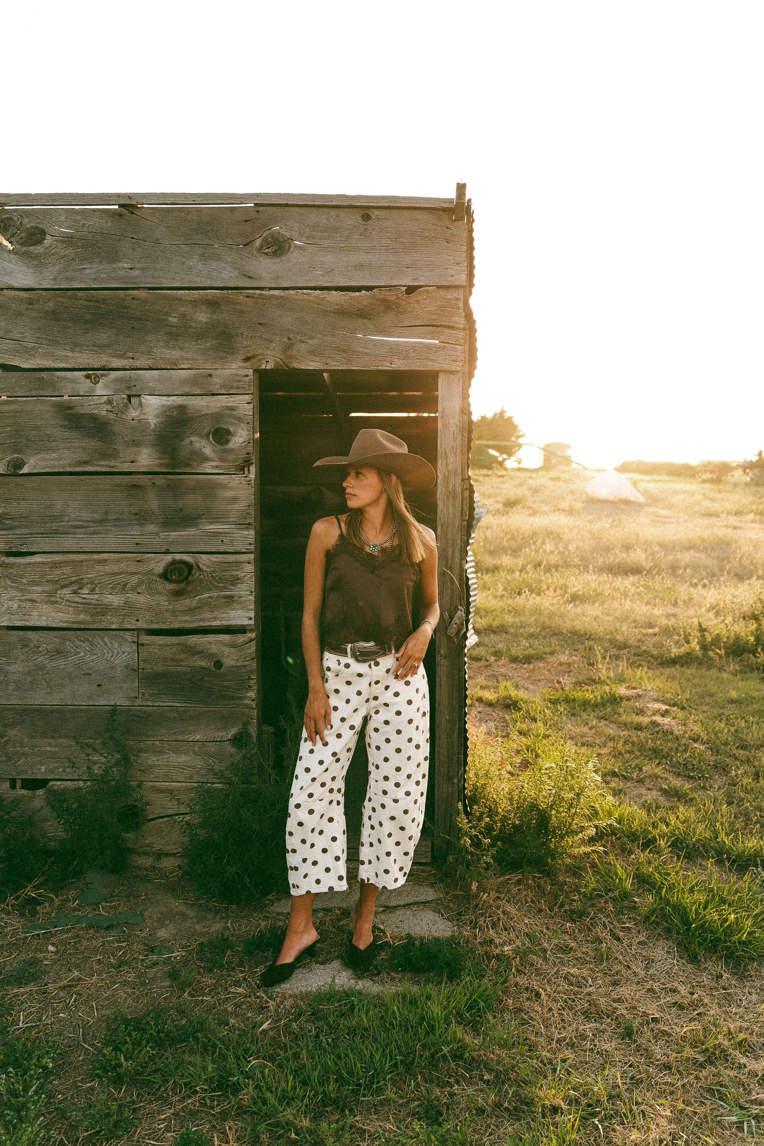 Woman in cowboy hat, black top, and polka dot pants leaning against rustic wooden barn at golden hour.