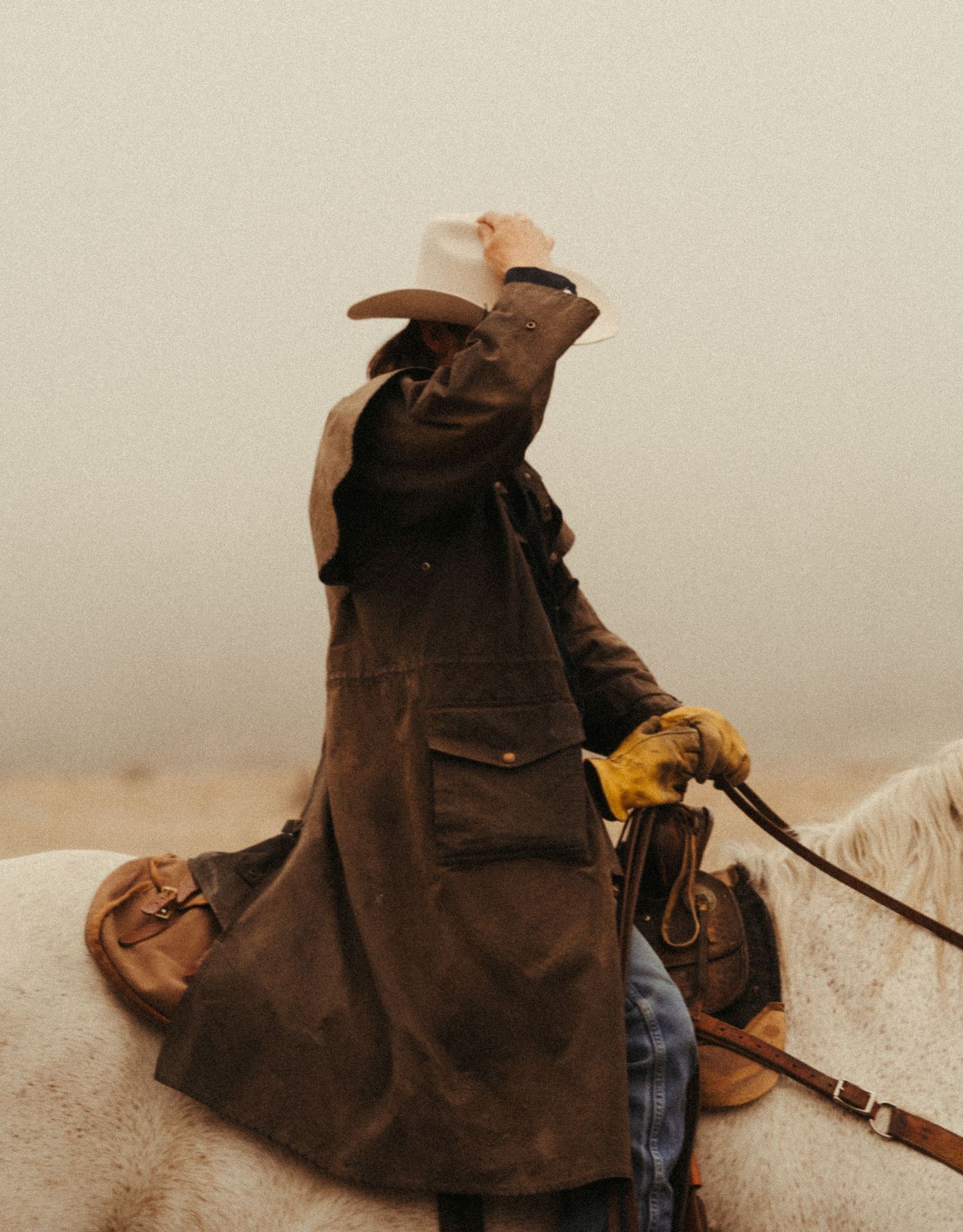 Cowboy in long brown duster coat on white horse, tipping white hat in misty conditions.