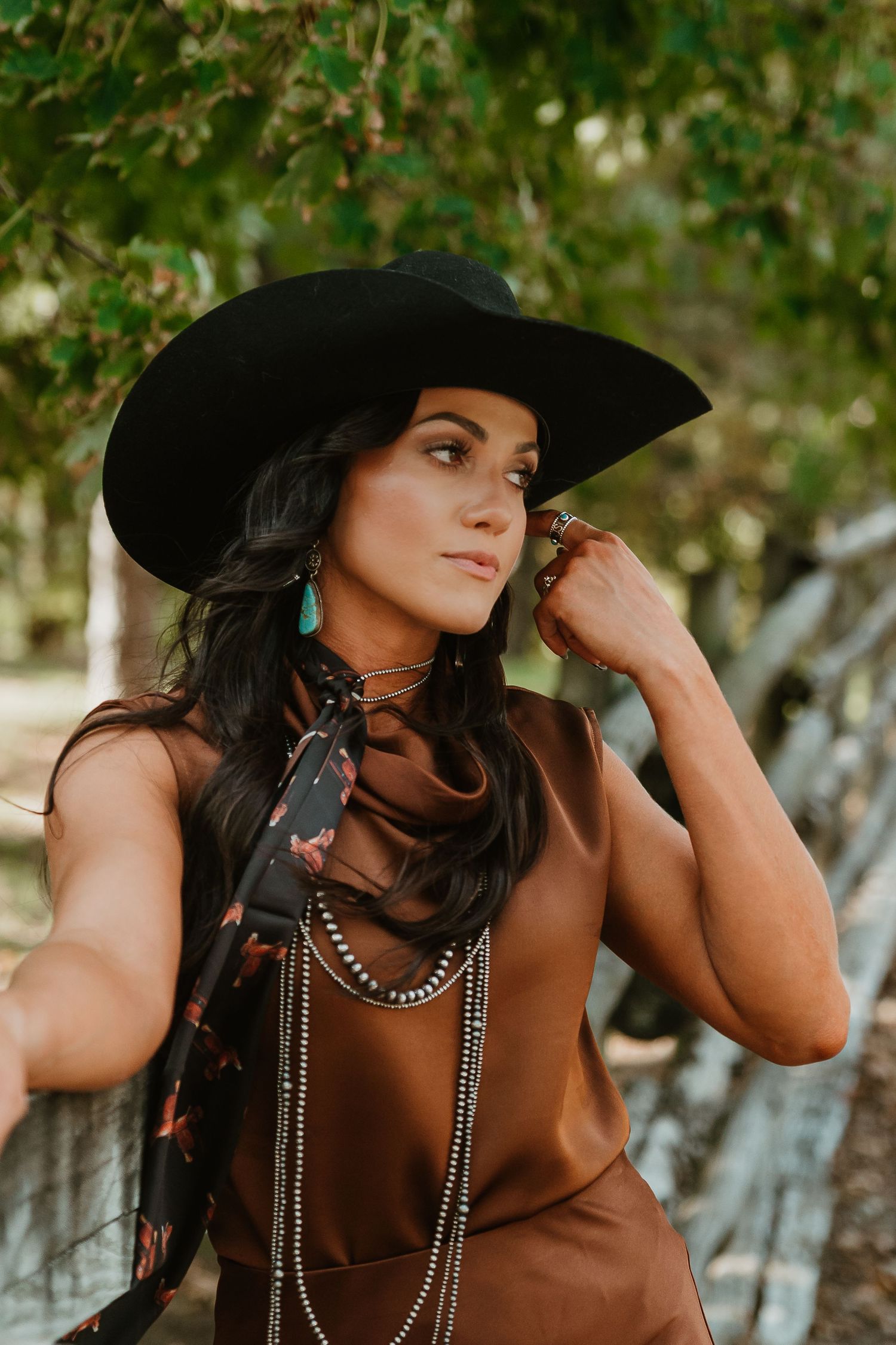 Woman in black wide-brim hat, brown satin top, turquoise earrings, and layered necklaces poses outdoors near a wooden fence.