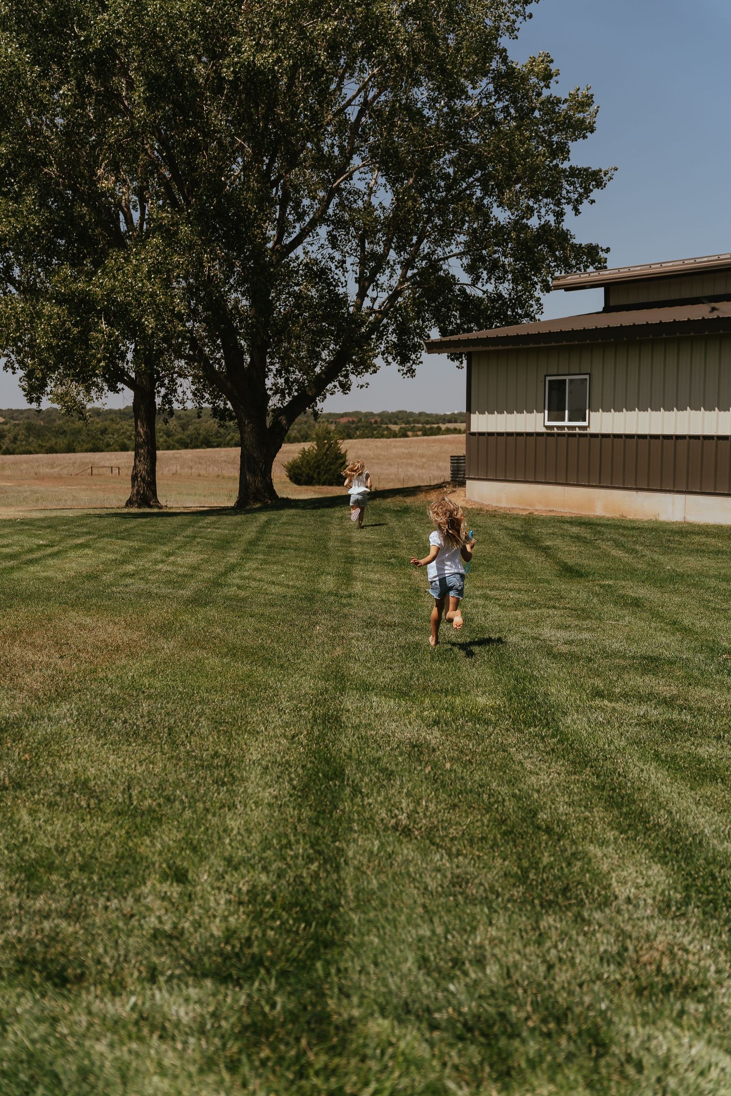 Two children running across a mowed green lawn beside a modern barn building with a large tree in the background.