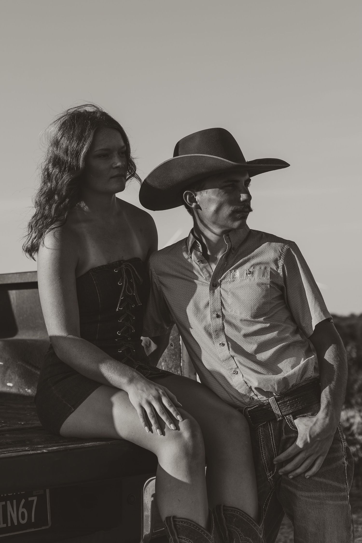 Black and white photo of a cowboy and young woman leaning against a vintage truck in a rural setting.