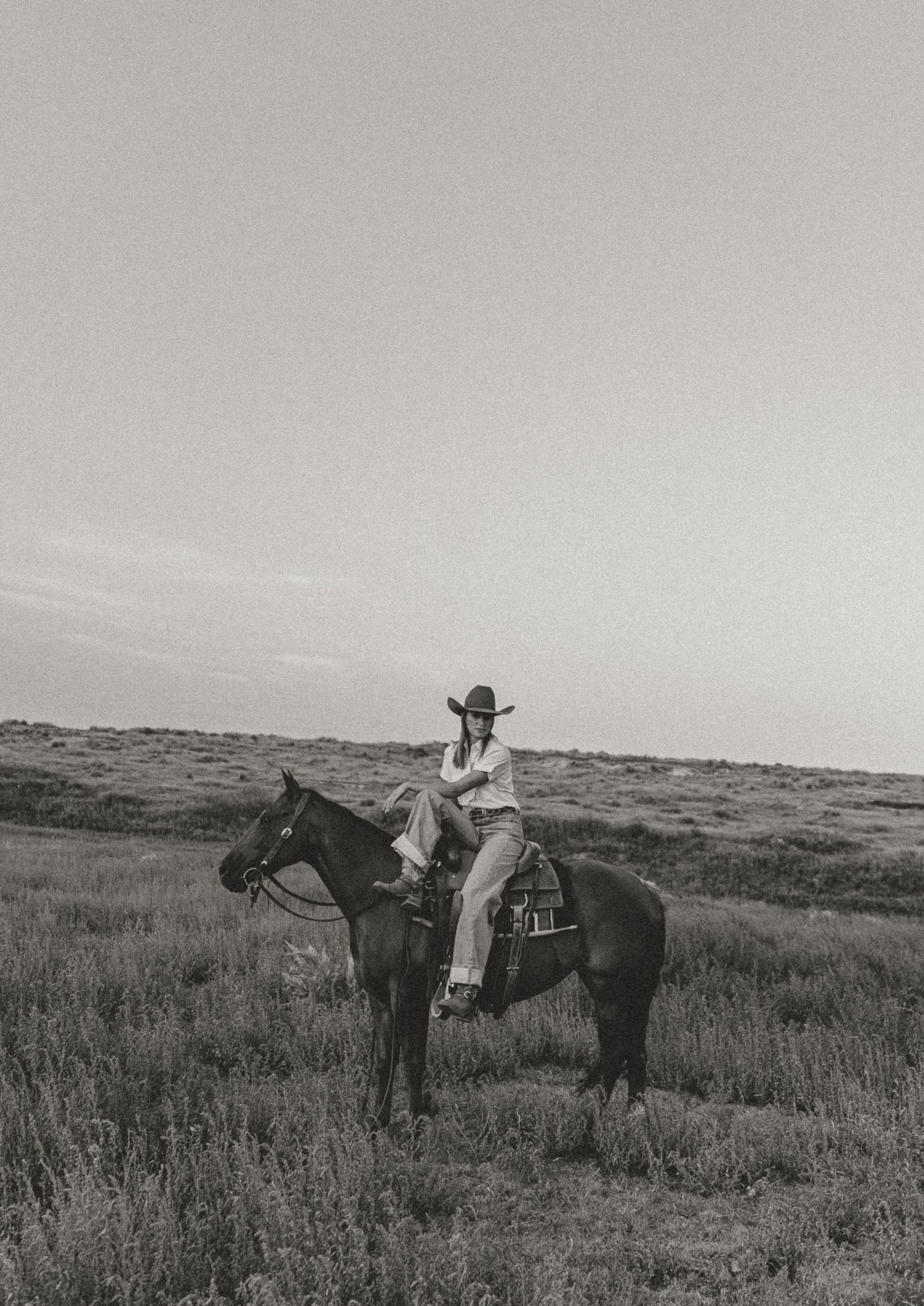 Black and white photo of a cowgirl in a hat riding a dark horse through an open grassy field.