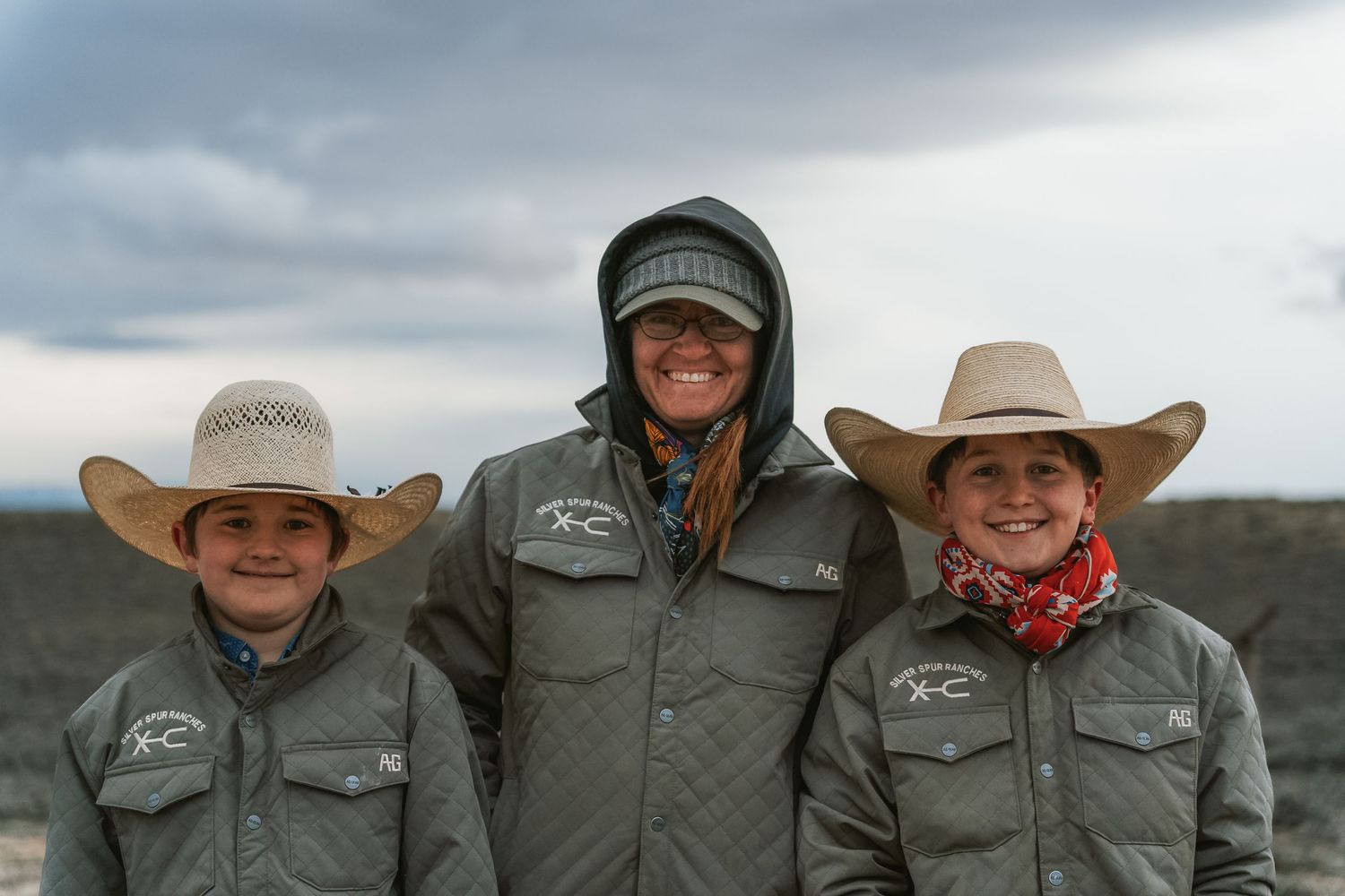 Three people in cowboy hats and matching gray jackets smile outdoors under a cloudy sky.