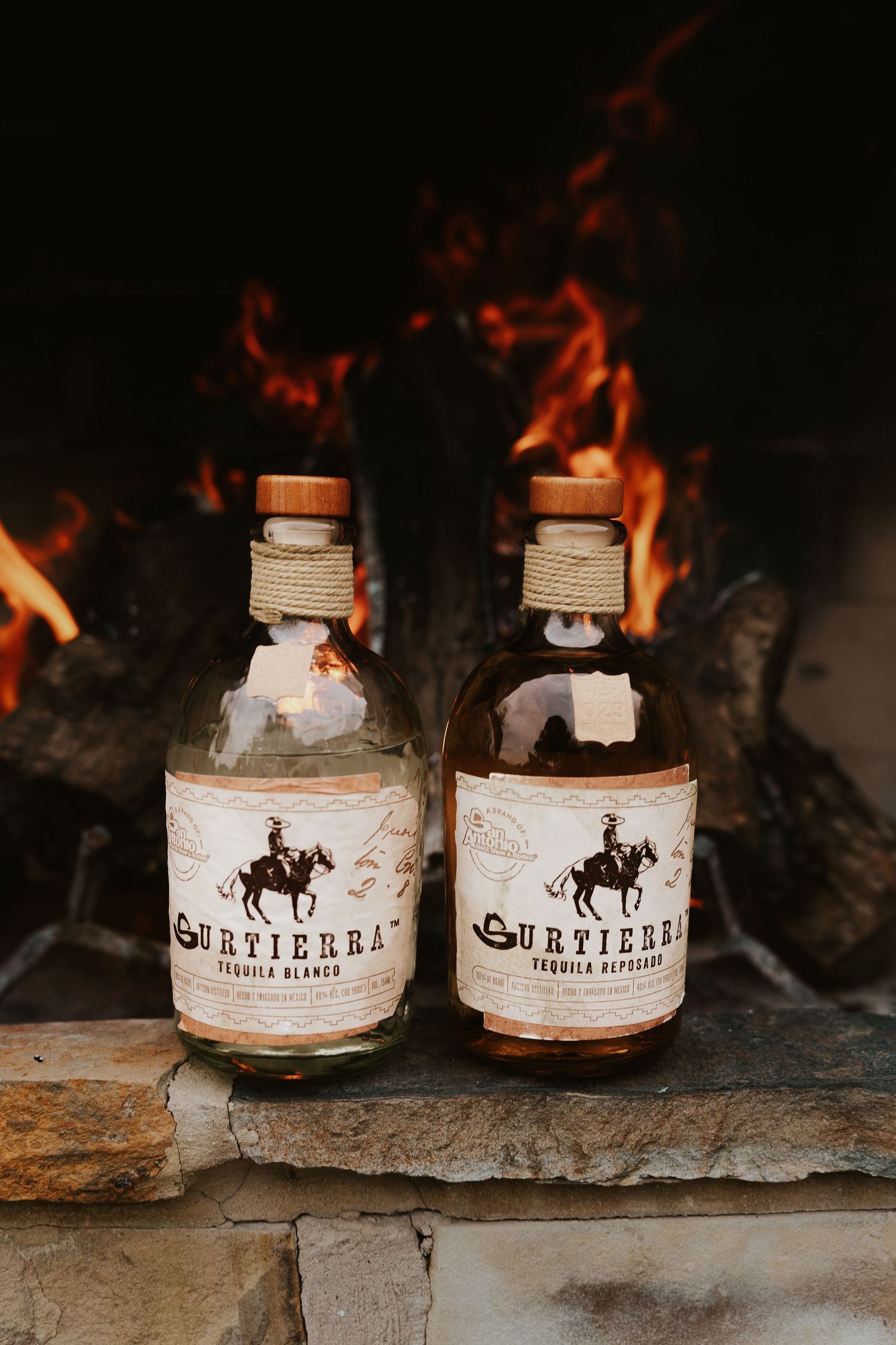 Two bottles of Surtierra Tequila Blanco and Reposado sit before a glowing fireplace on stone hearth.