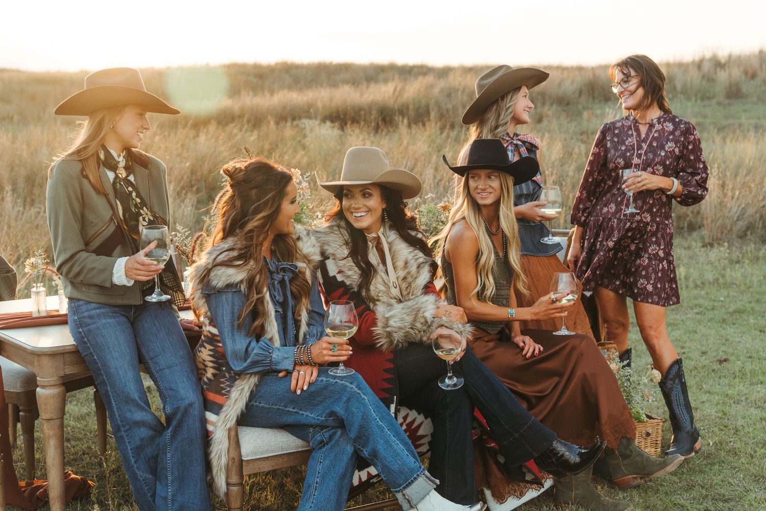 Women in cowboy hats enjoying wine outdoors in a field at golden hour, dressed in western boho fashion.