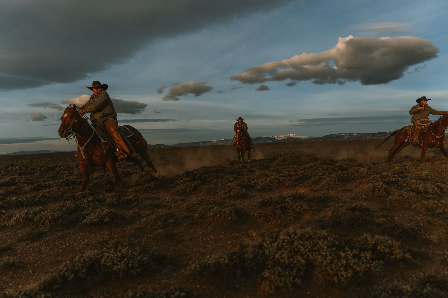 Three cowboys riding horses across a dusty ridge under a dramatic cloudy sky at dusk.