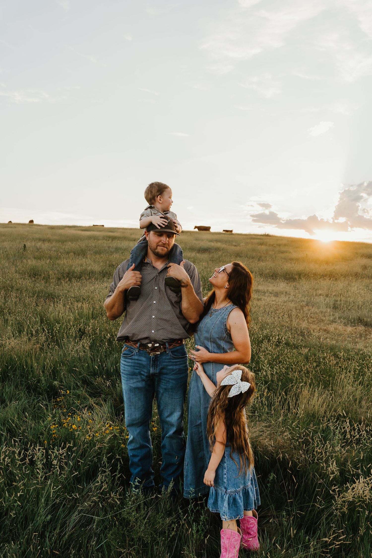 Family posing in golden sunset field, father carrying toddler on shoulders, mother and daughter in denim dresses.
