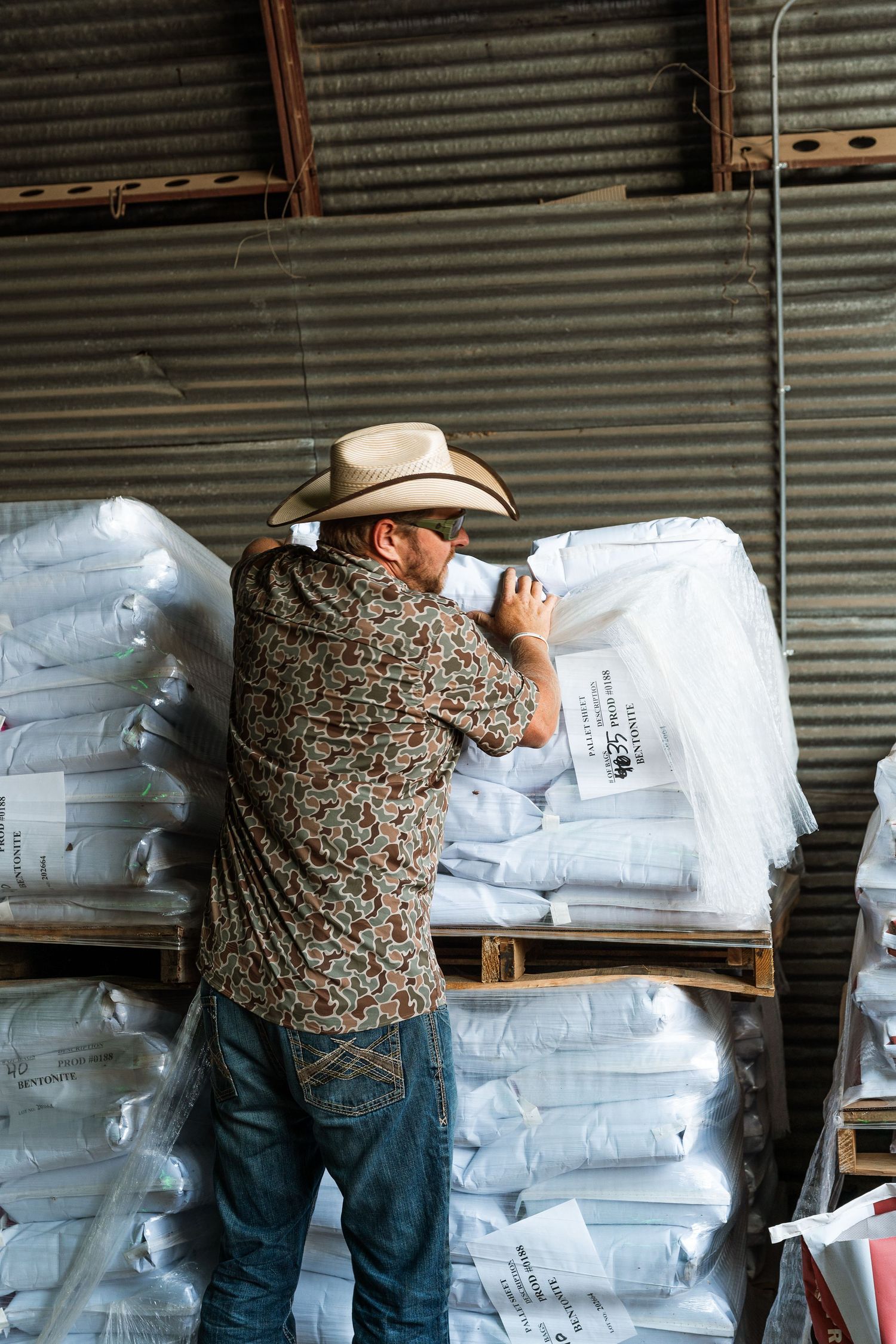 Man in cowboy hat and floral shirt examining white bags on warehouse pallet.