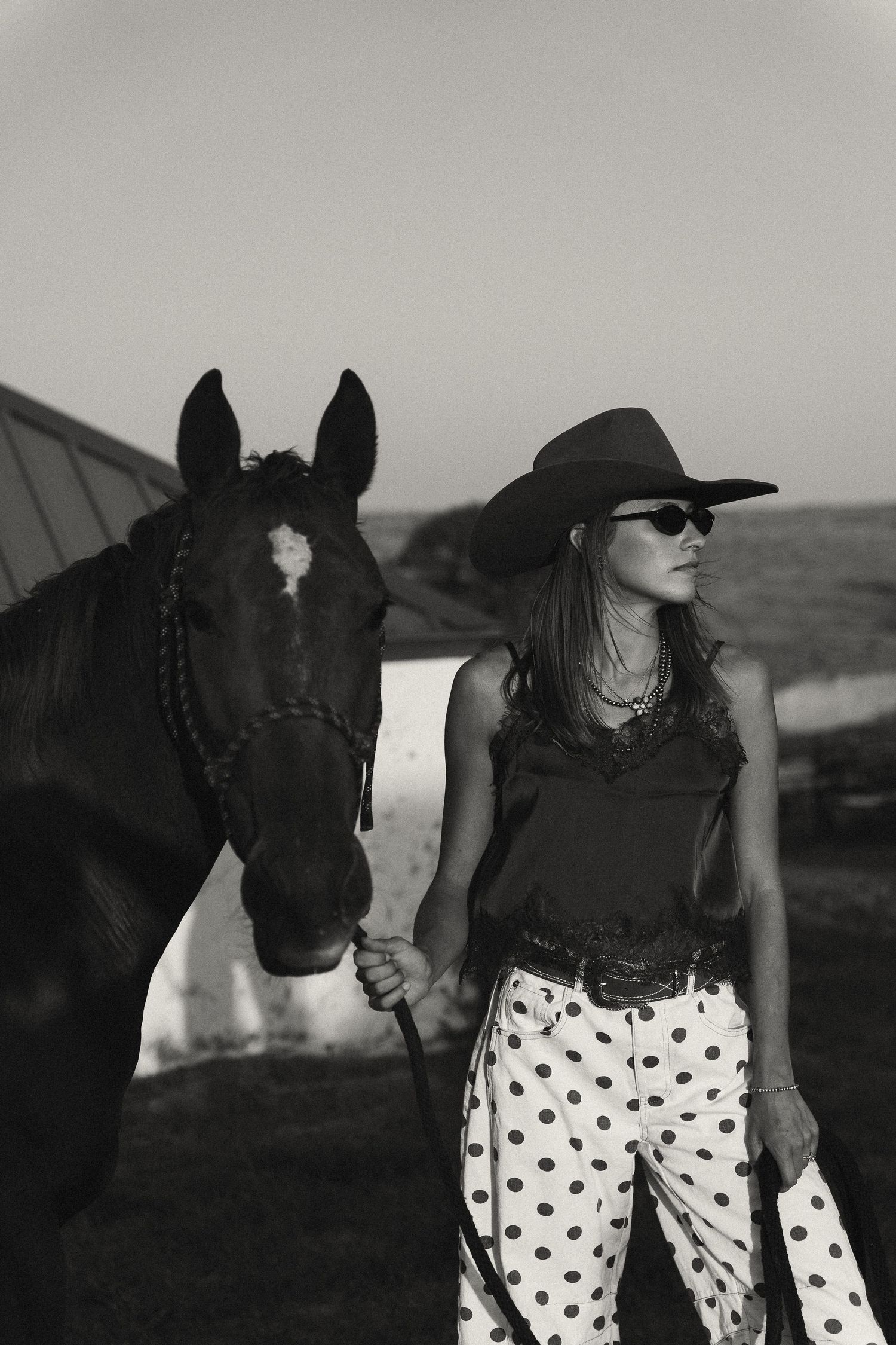 Black and white photo of a cowgirl in polka dot pants and hat standing beside a dark horse near a barn.