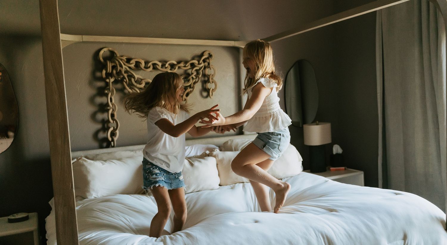Two girls jumping and playing on a white hotel bed in a cozy room with decorative headboard.