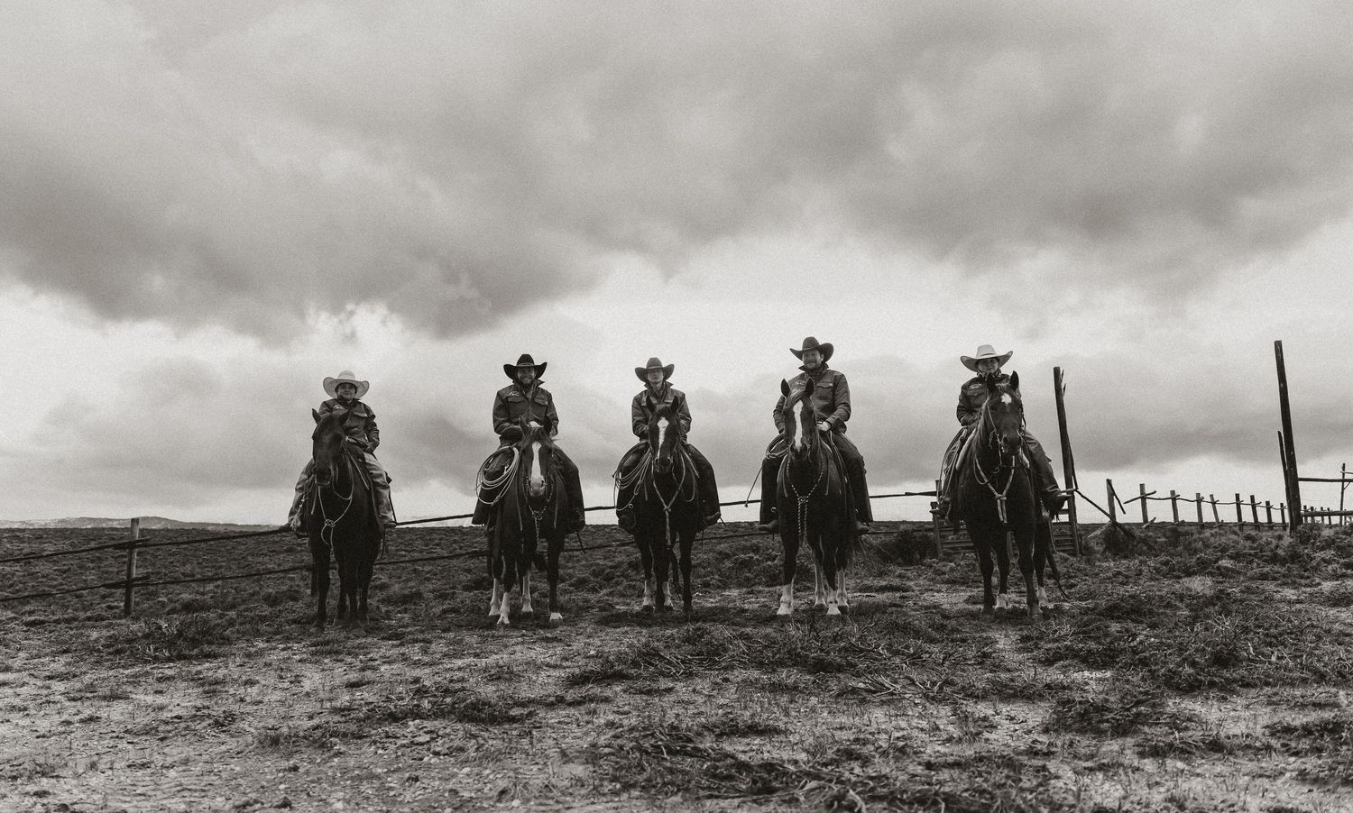 Five cowboys on horseback ride across an open plain under dramatic cloudy skies in a classic black and white photo.