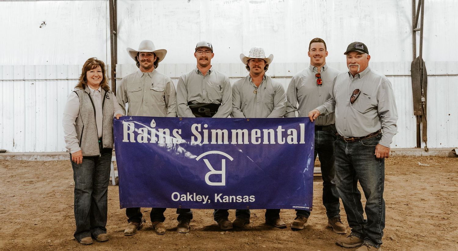 Group photo of Rains Simmental cattle ranch team holding banner in Oakley, Kansas indoor arena.