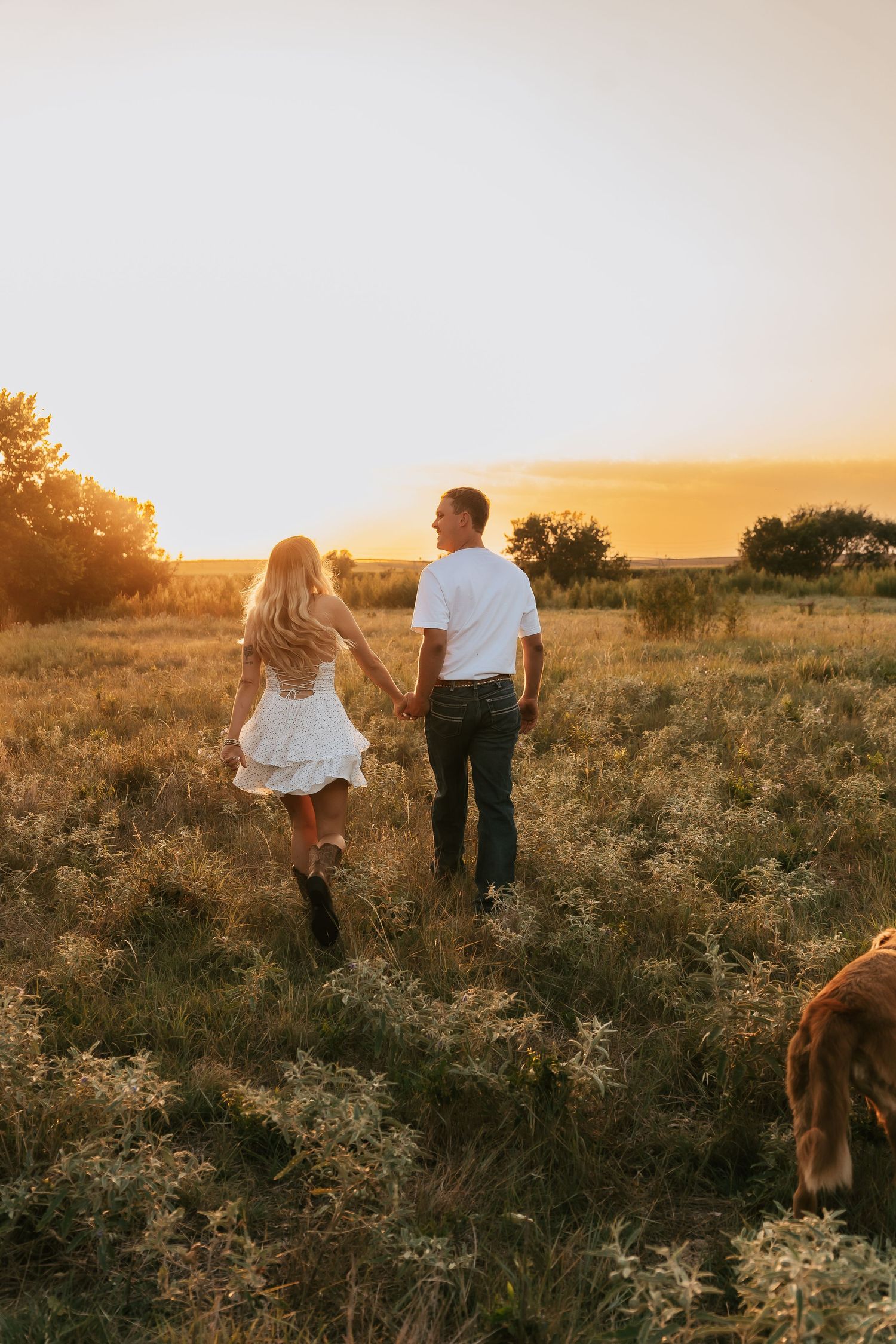 Couple holding hands walking through golden field at sunset, woman in white dress, man in white shirt and dark jeans.