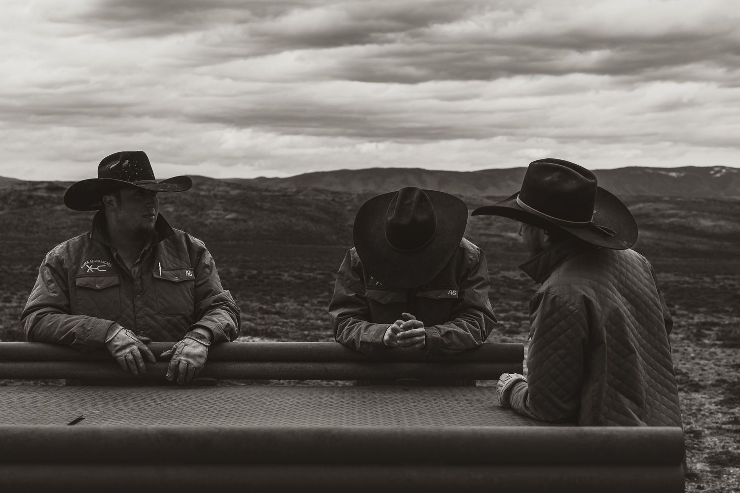Three cowboys in black hats sit on a truck tailgate against a dramatic rural landscape in black and white.