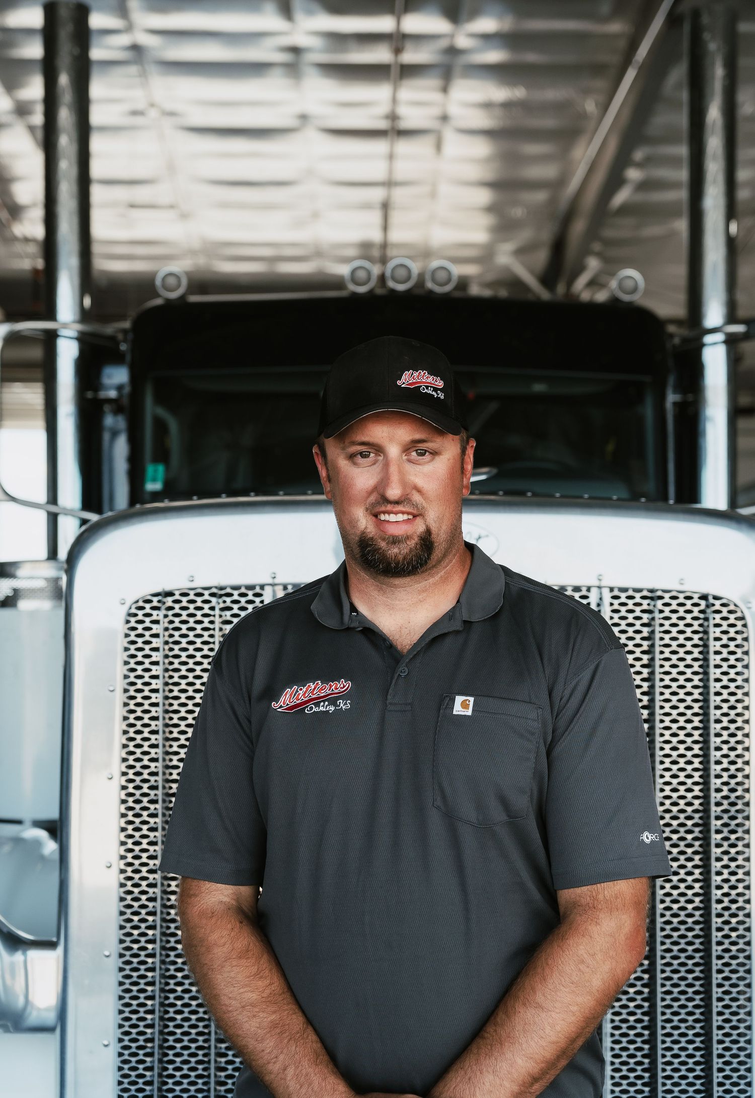 A smiling truck driver in a dark polo shirt and cap stands confidently in front of a large semi-truck.
