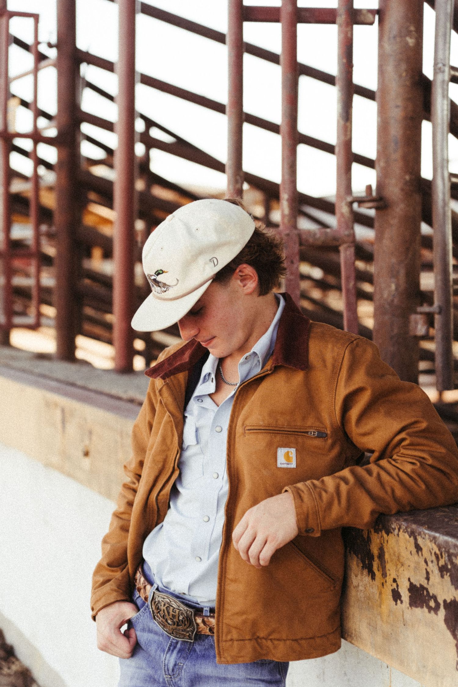 Young man in Carhartt jacket and white cap leaning against a rustic metal fence at a rodeo arena.