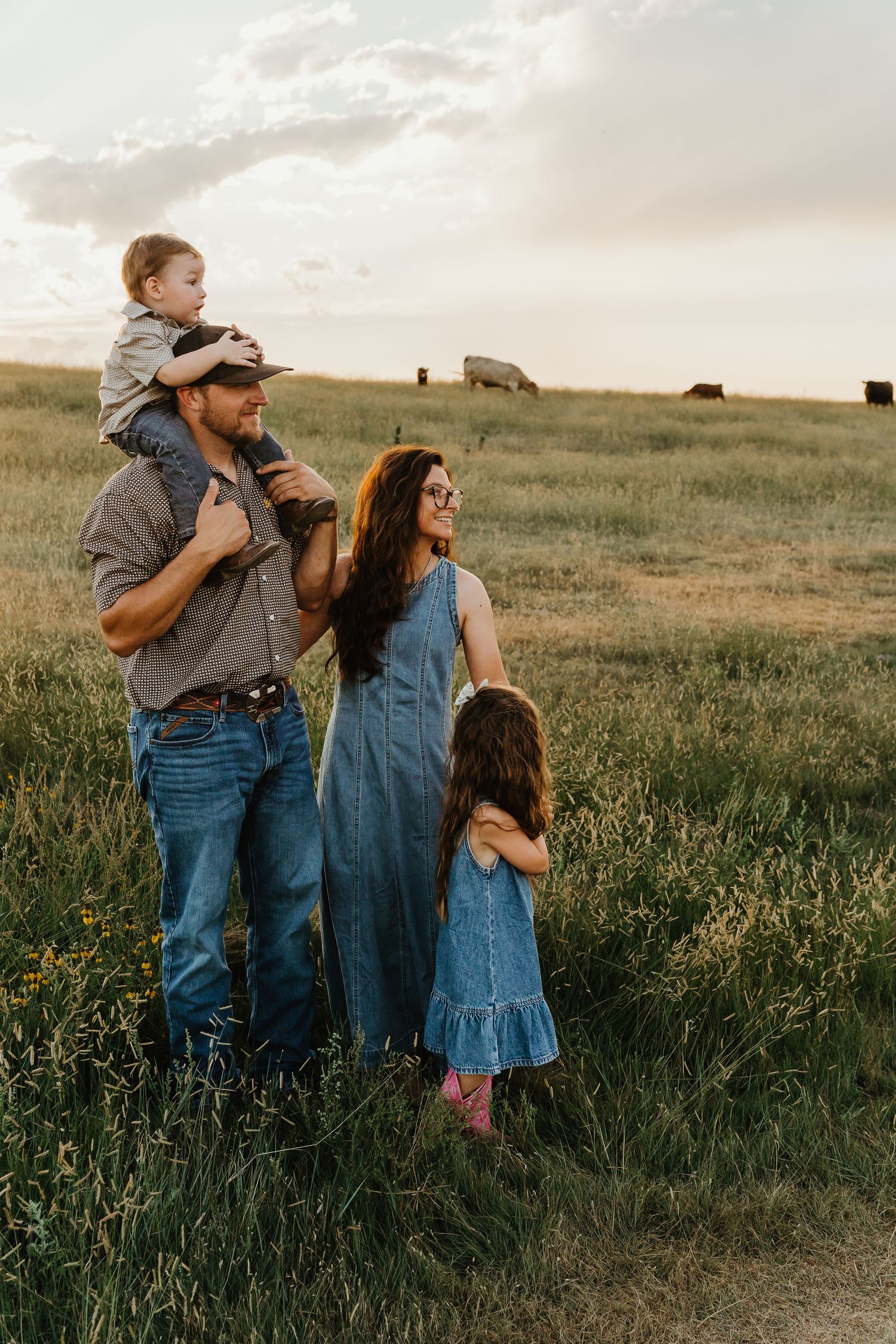 Family of four in denim outfits standing in green pasture at golden hour with cattle grazing.