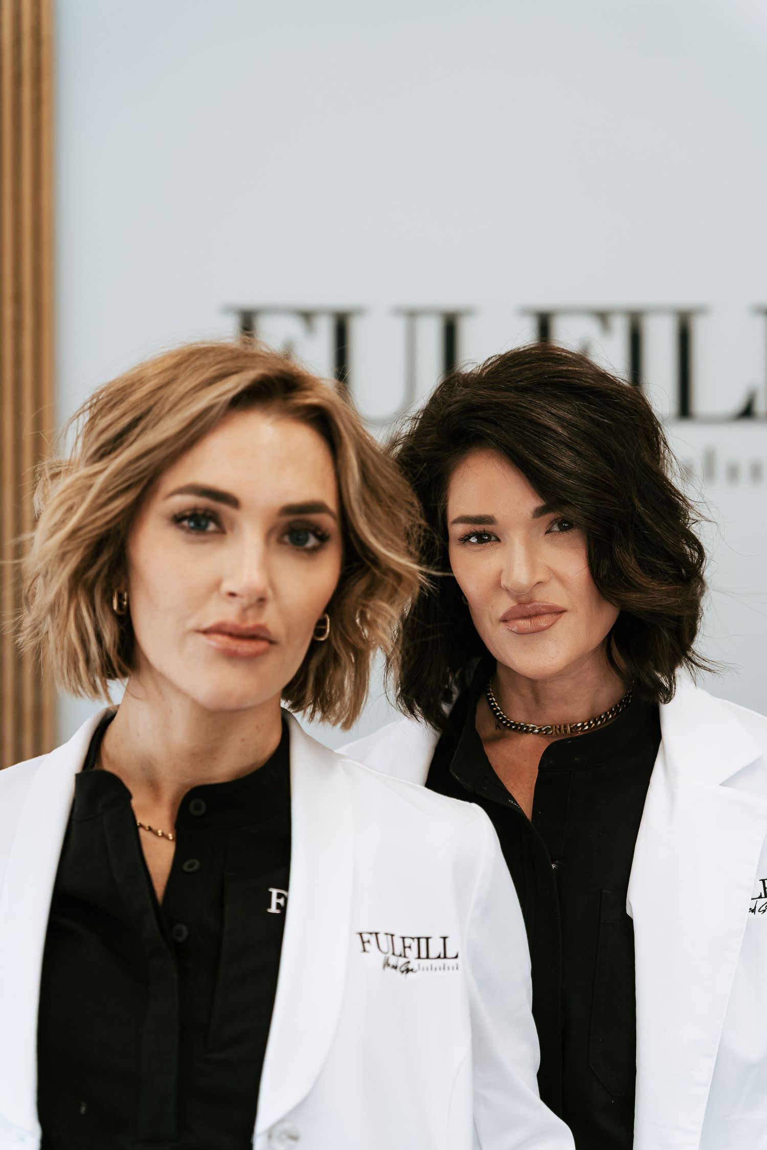 Two women in white Fulfill lab coats pose professionally in front of a Fulfill branded backdrop.