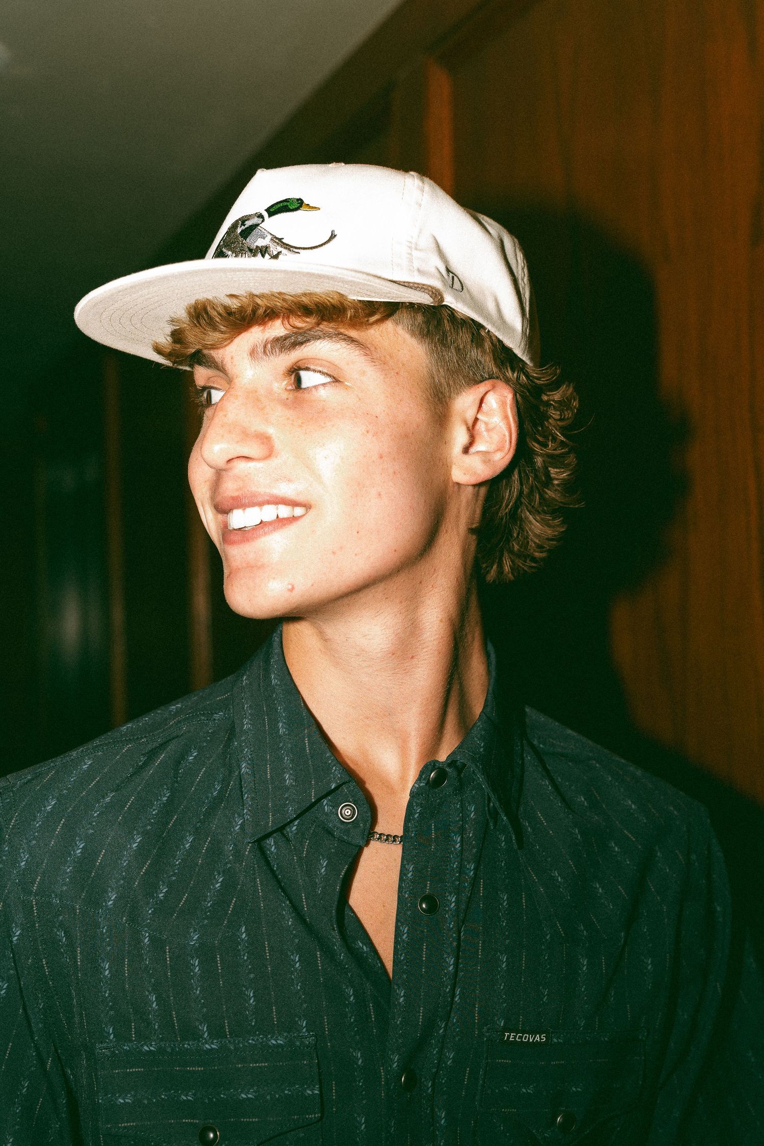 Young man smiling in a white snapback cap and dark pinstripe shirt, looking sideways indoors.
