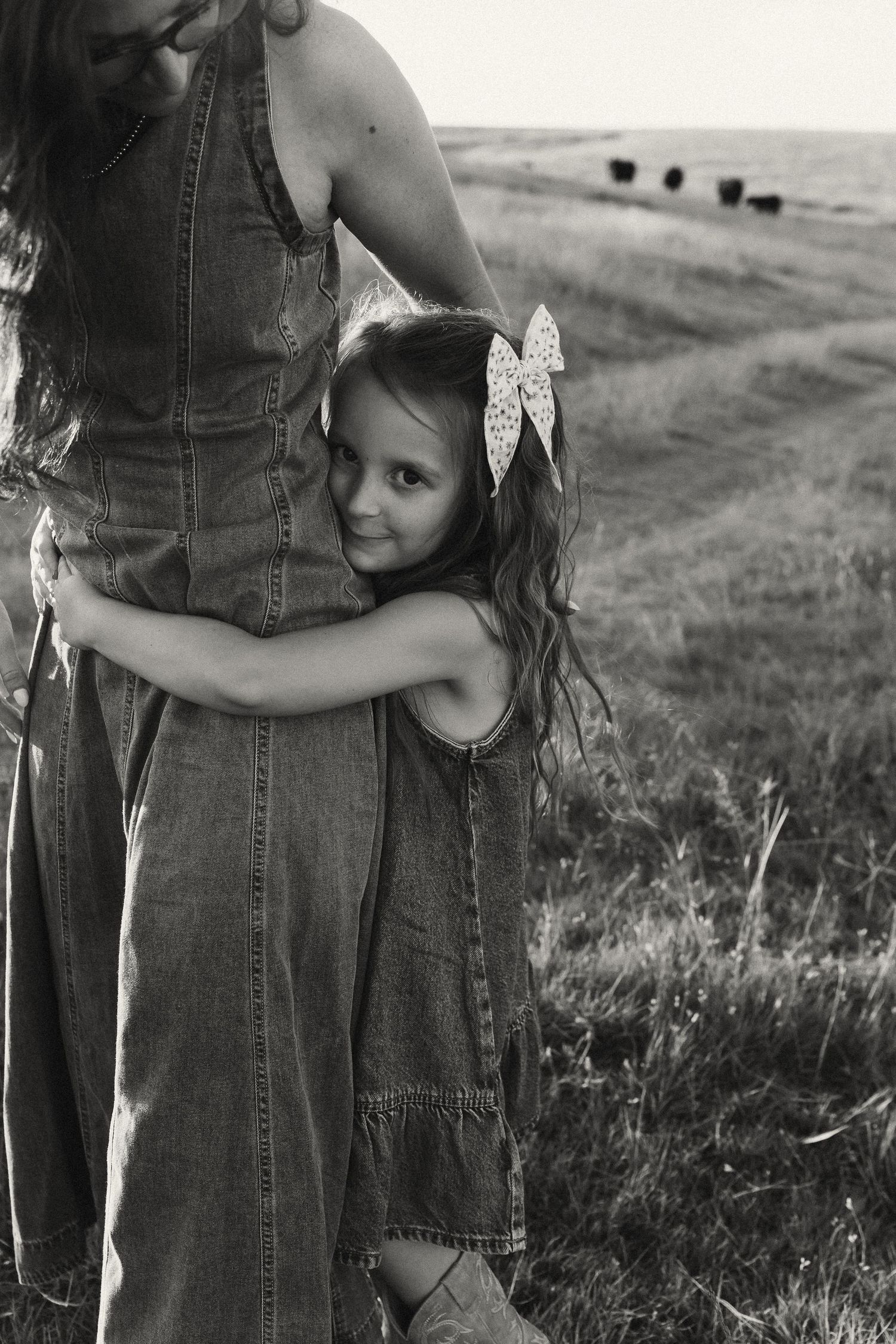 Black and white photo of young girl hugging mothers legs in grassy field with cattle.