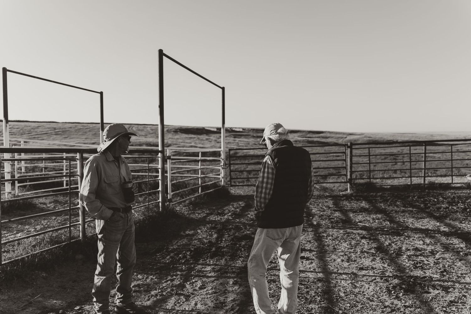 Two farmers discuss near metal cattle gates on an open prairie ranch in a black and white photo.