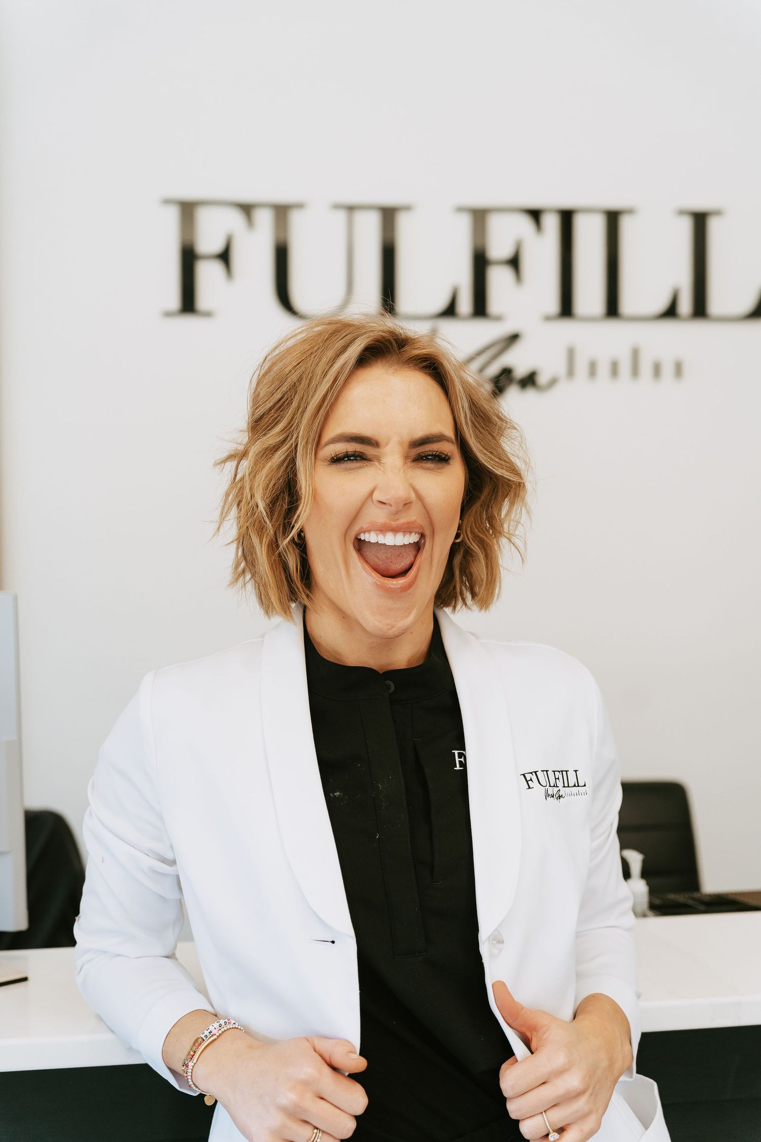 Smiling woman in white blazer and black shirt stands at Fulfill salon reception desk.