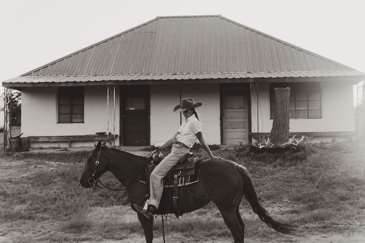 Black and white photo of a cowboy riding a dark horse in front of a rural ranch house.
