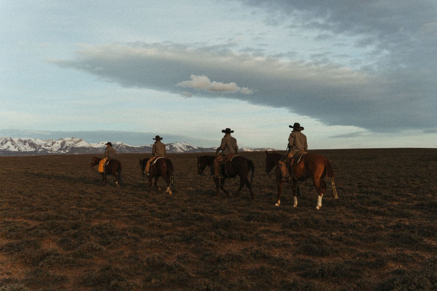 Three cowboys on horseback ride across a vast open plain with snow-capped mountains under a dramatic sky.