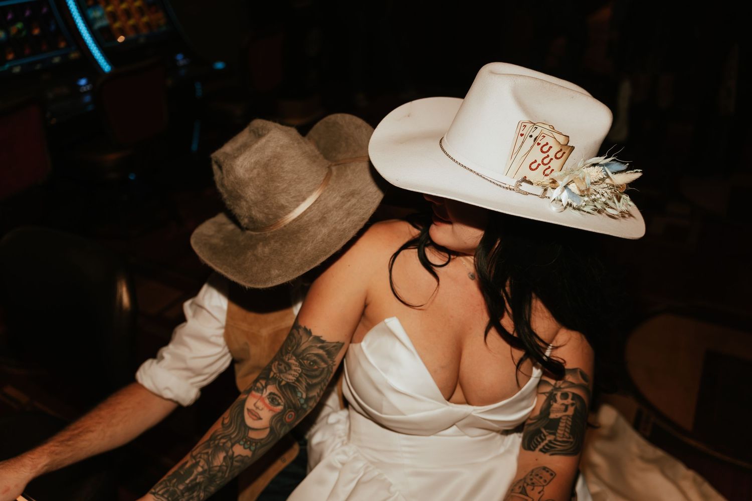 Tattooed couple in cowboy hats sharing a romantic moment, bride in white dress with decorated hat at wedding reception.