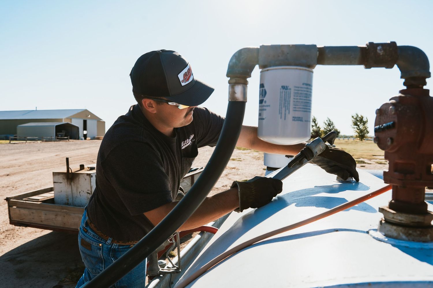Technician in black uniform and cap connecting fuel hose to agricultural equipment on a sunny farm.