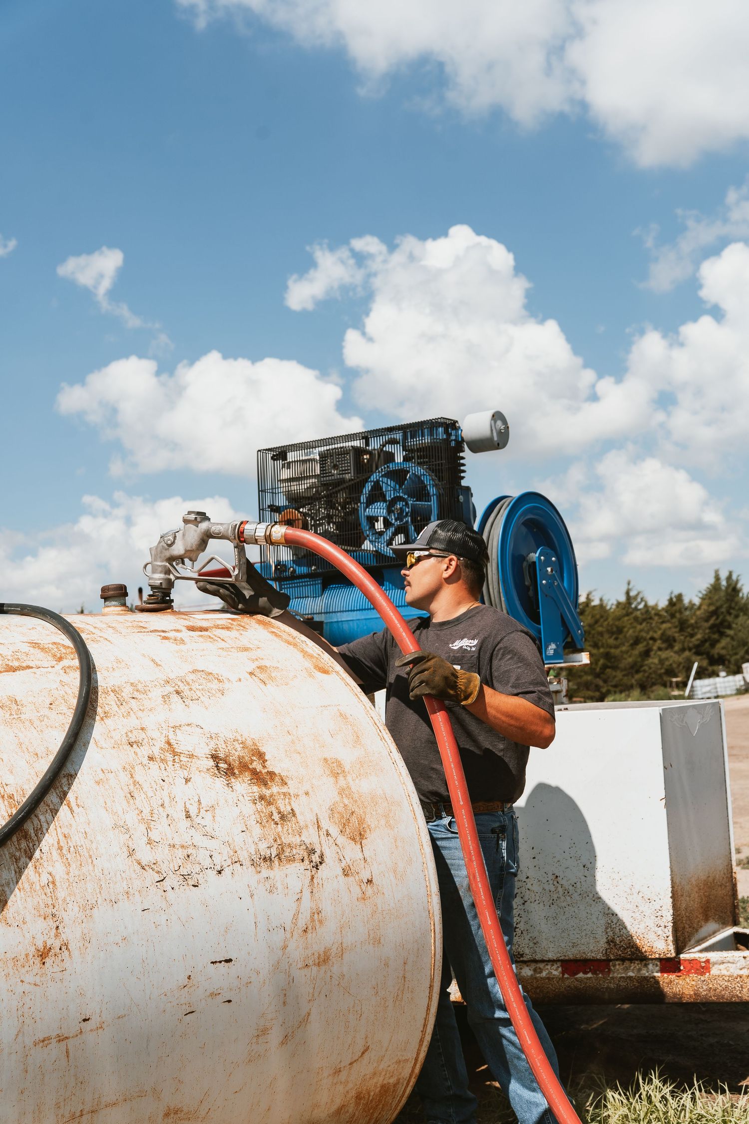 Worker connects red hose to rusty fuel tank outdoors with blue pump equipment and cloudy sky in background.