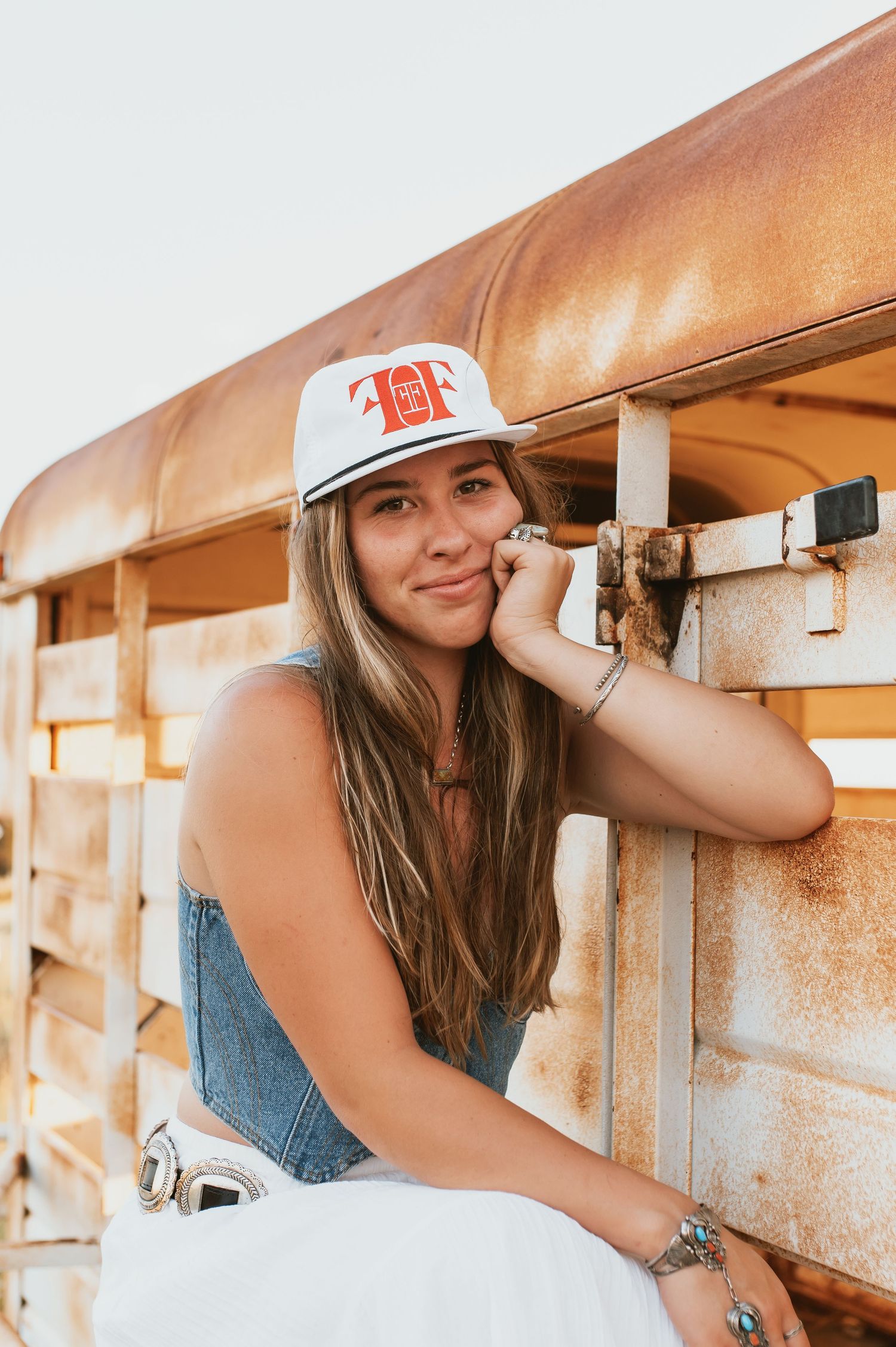 Young woman in denim overalls and white snapback cap leans against rustic metal fence outdoors.
