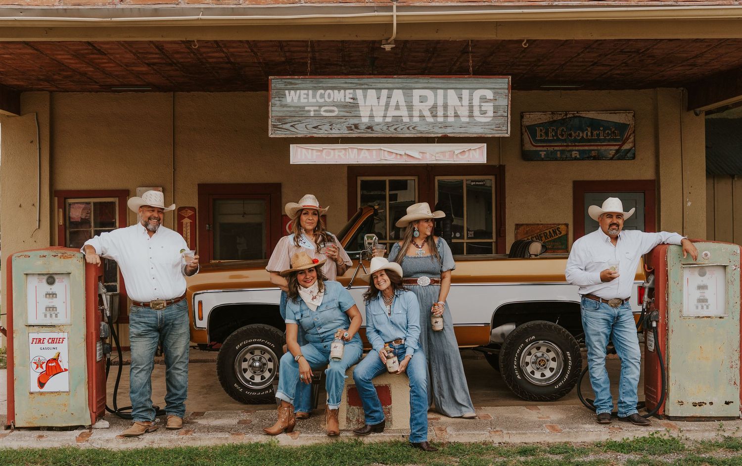 Country group poses in cowboy hats near a Welcome to Waring sign beside a vintage truck at a rustic Texas general store.